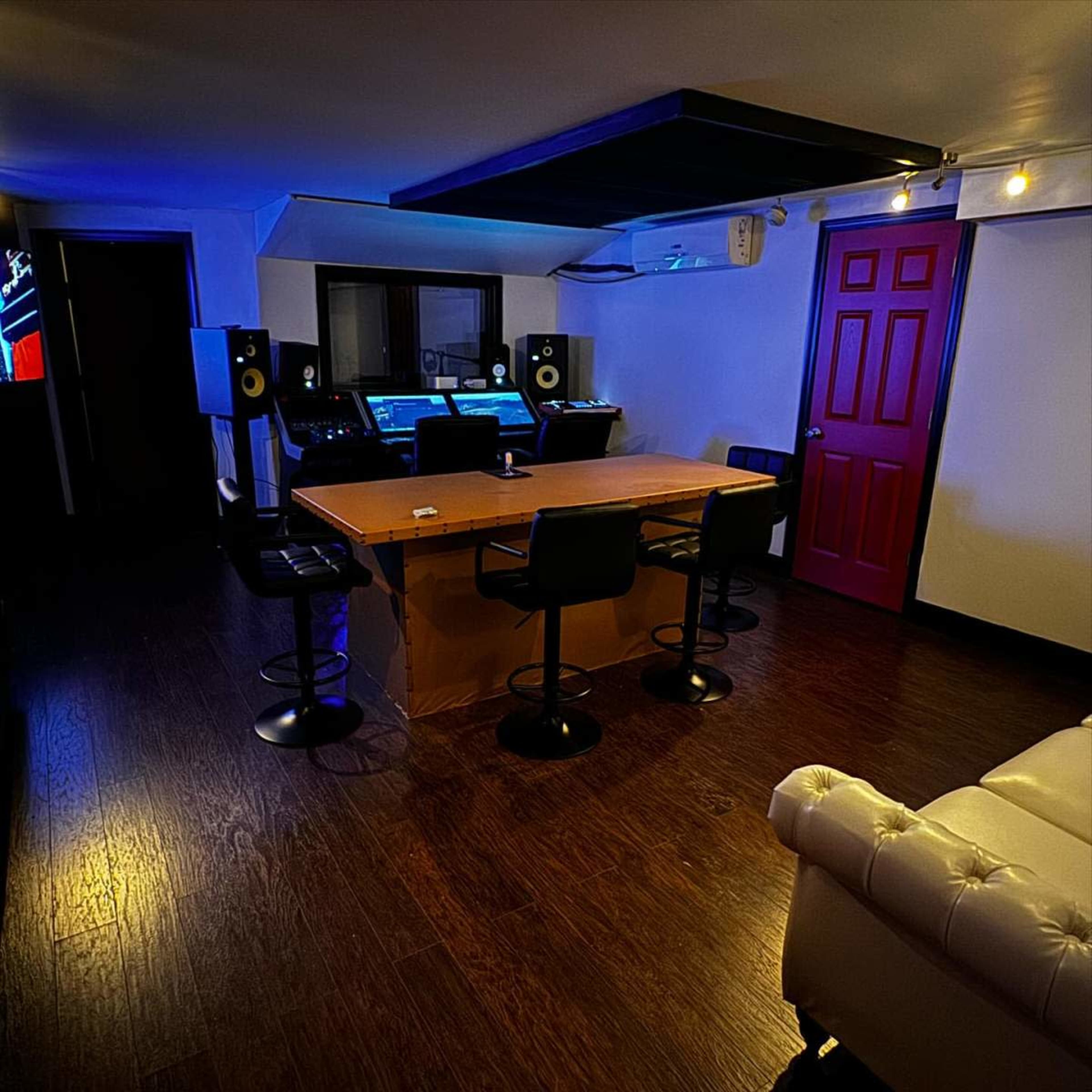 The image shows a music studio with a central table surrounded by four stools, two large speaker monitors, and a computer setup against a backdrop featuring a red door and ambient blue lighting.
