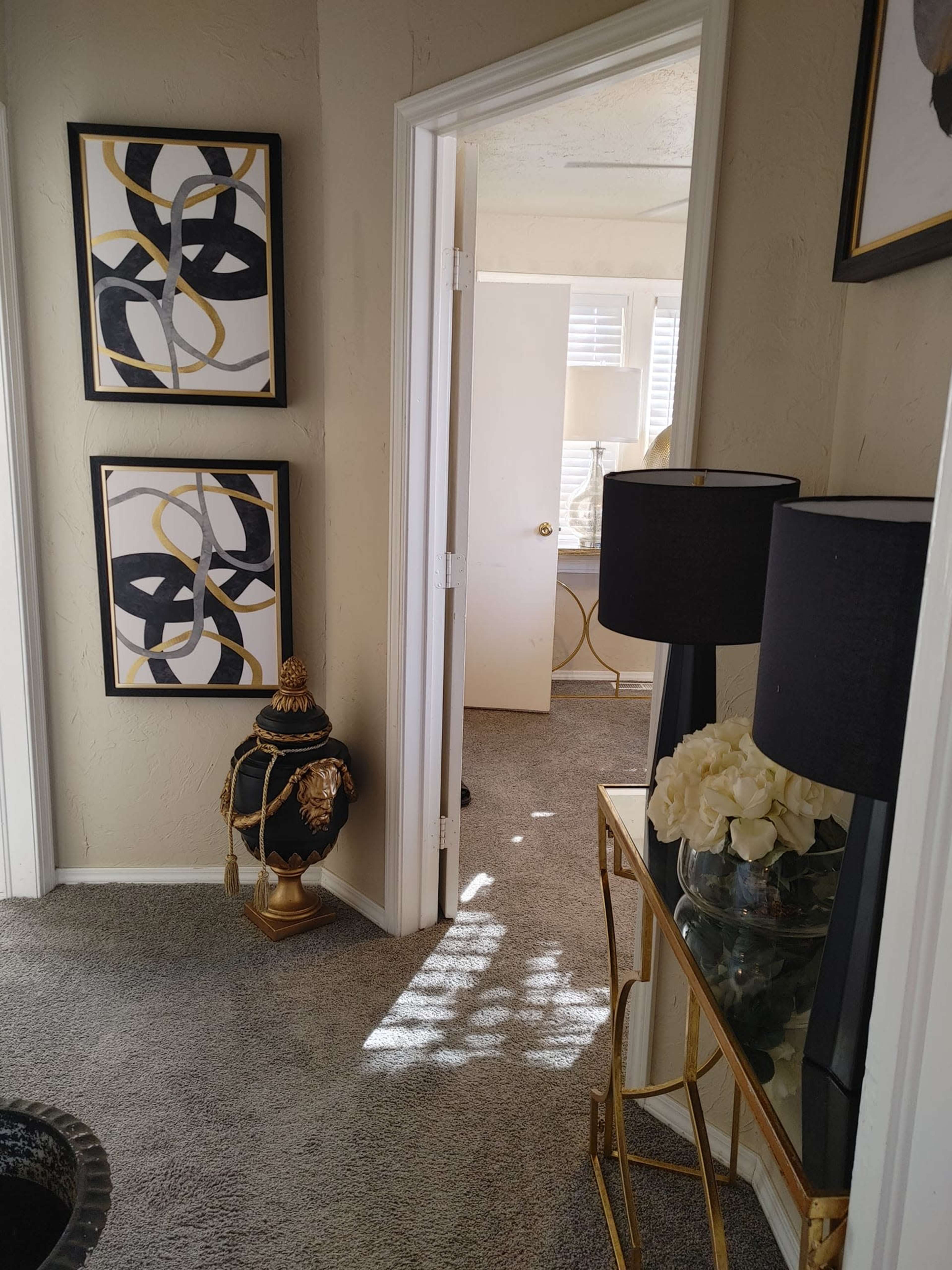 A well-lit hallway leading to an adjacent room, with abstract framed art on the walls and a decorative lamp and table in the foreground.