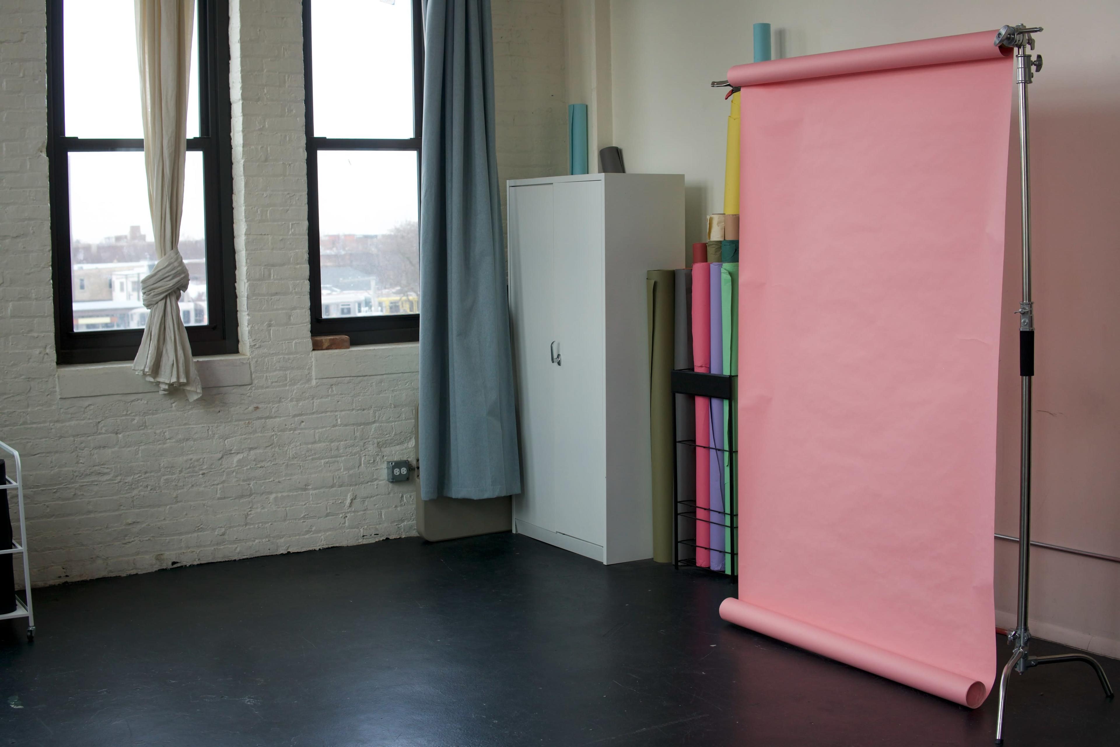 The image shows a studio space with a pink backdrop on a stand, a cabinet, and rolls of colorful paper stored nearby.