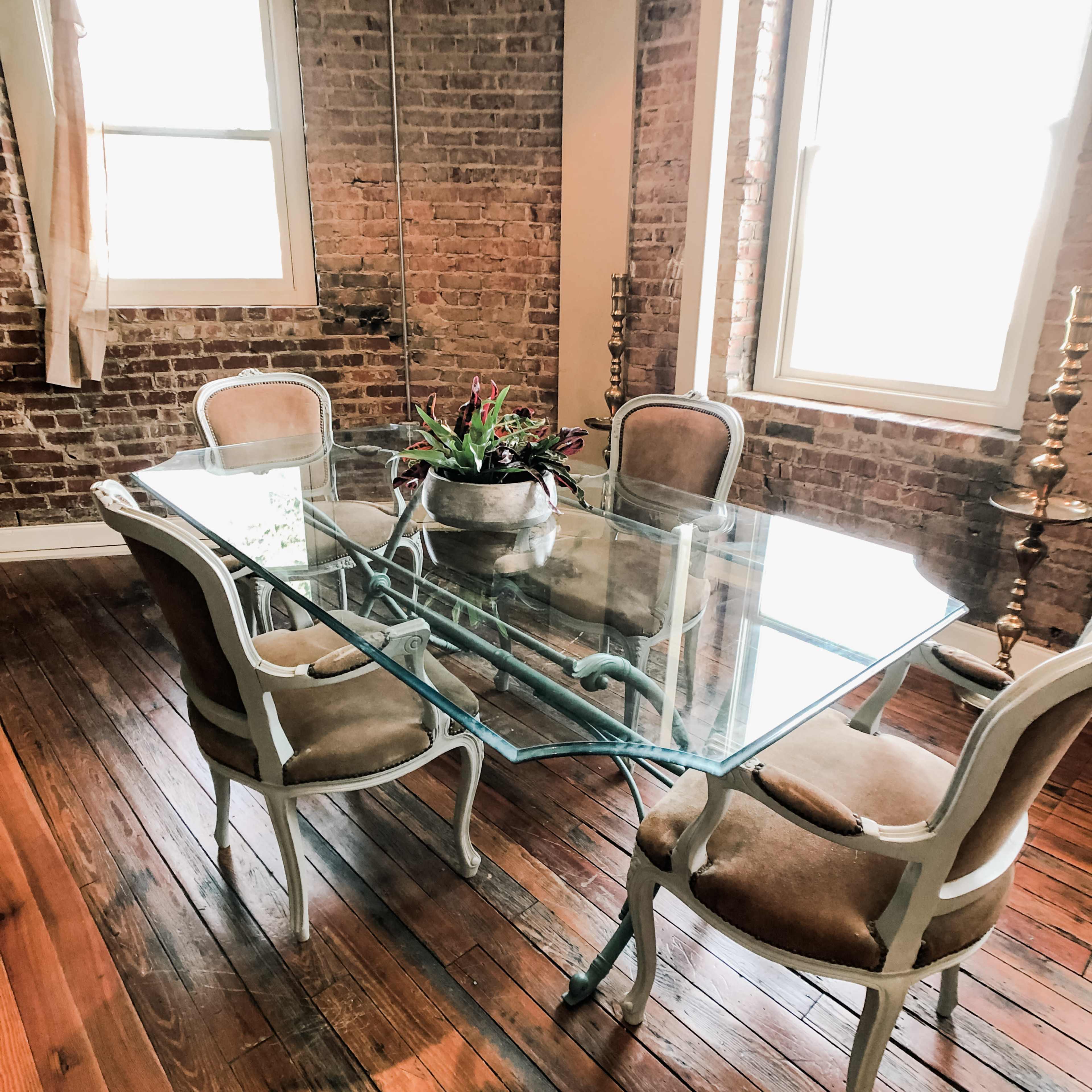 A glass-top table surrounded by four upholstered chairs is set in a room with exposed brick walls and wooden flooring.