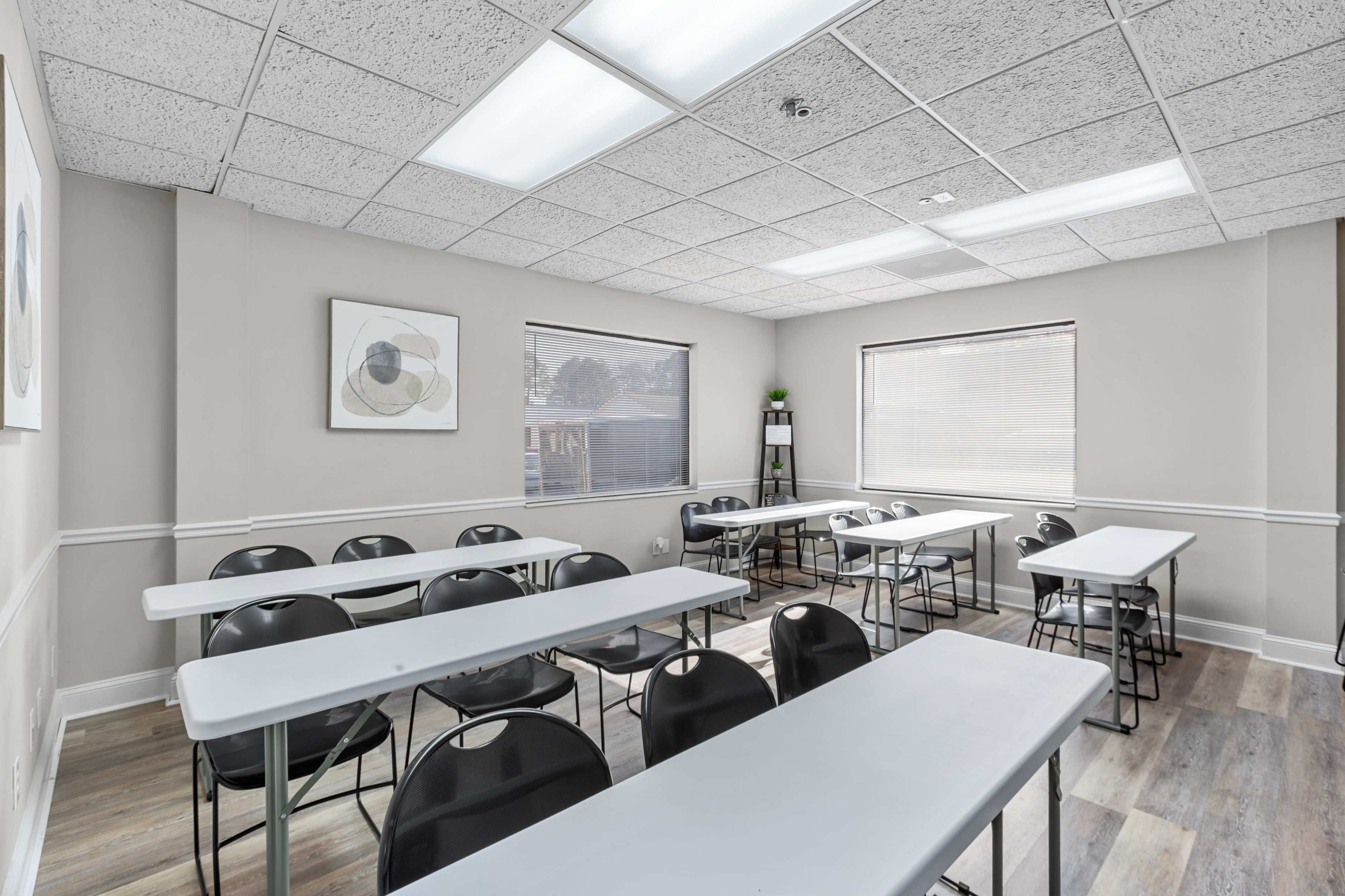 A classroom is set up with rows of tables and chairs, featuring large windows and a neutral-colored wall.