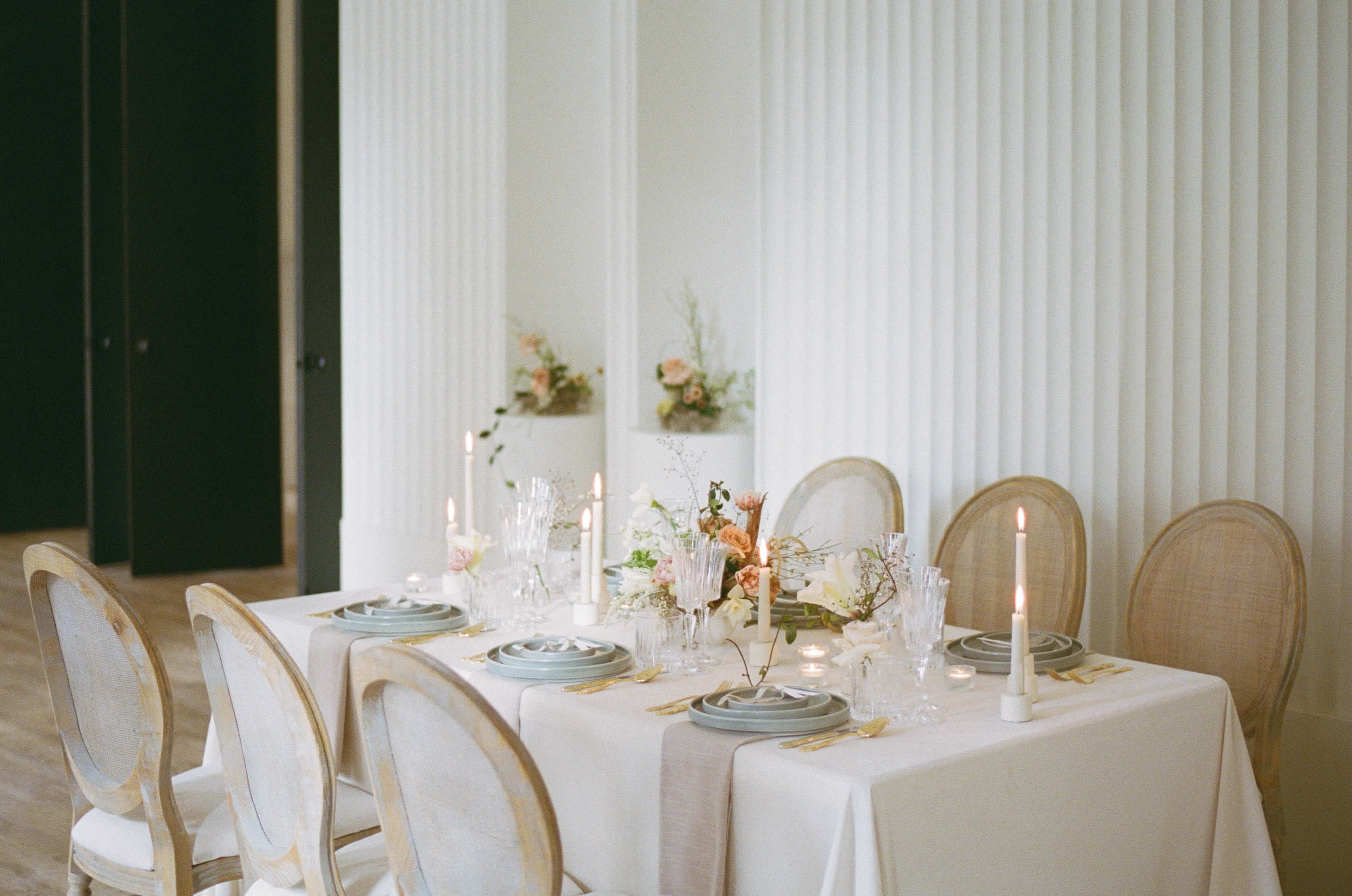 A dining table is elegantly set with candles and floral arrangements against a backdrop of textured white walls.