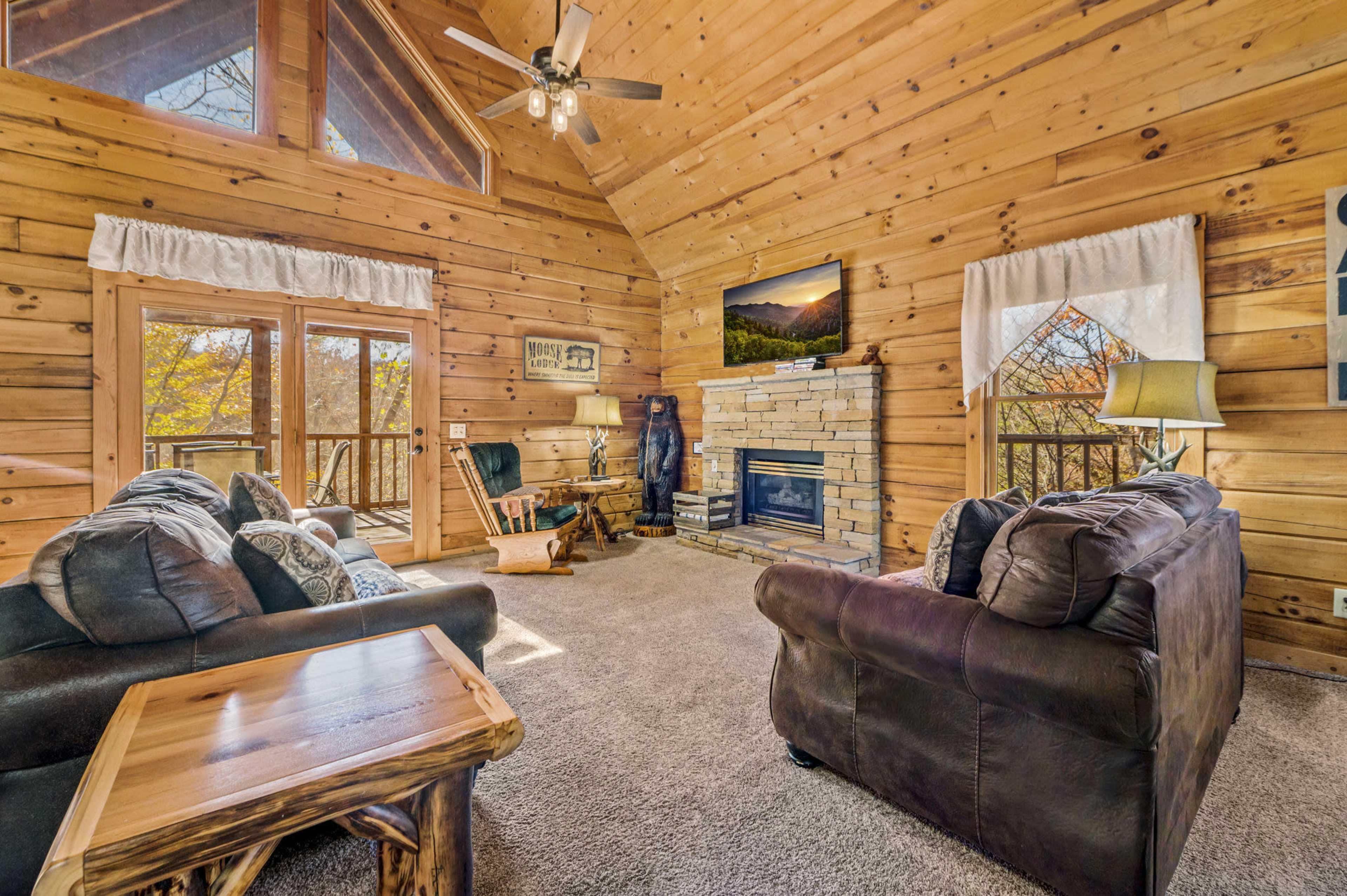 A cozy, wooden-paneled living room with a stone fireplace, large windows, and two dark leather sofas.