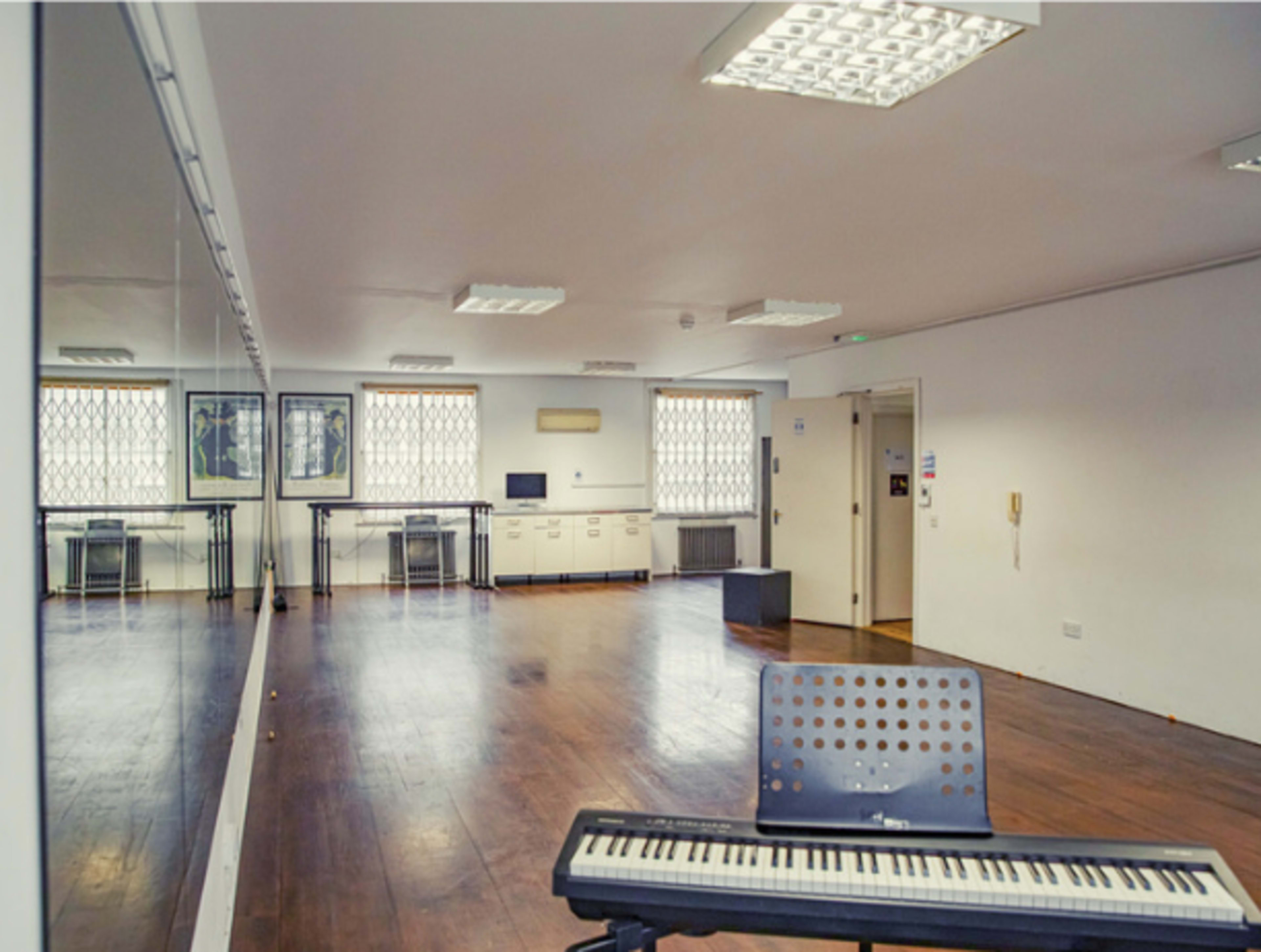 An empty room with wooden flooring, a piano in the foreground, and large windows along one wall reflecting in a mirror.