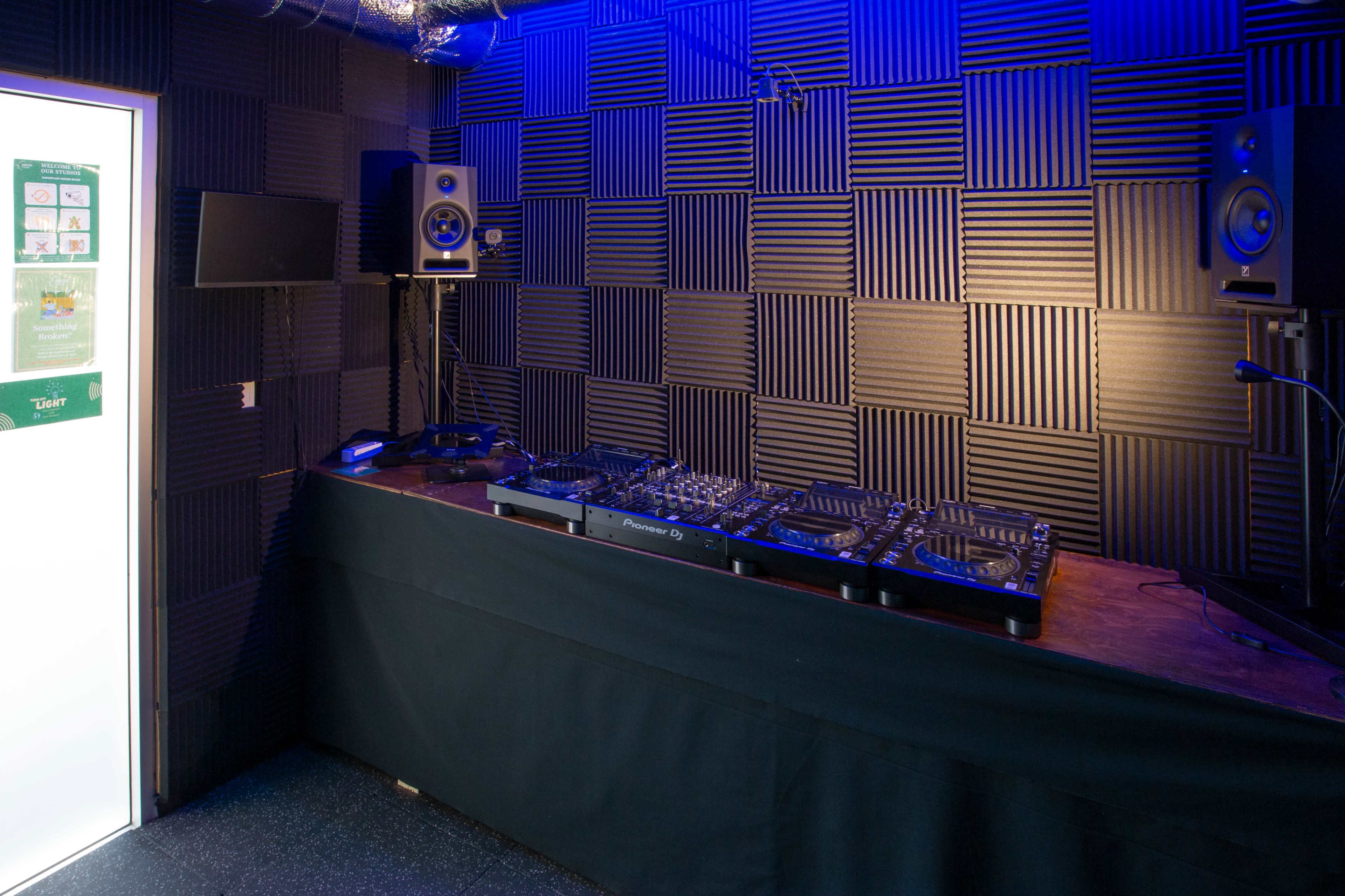A soundproof music studio features a DJ booth with equipment and speakers against a textured, dark wall.