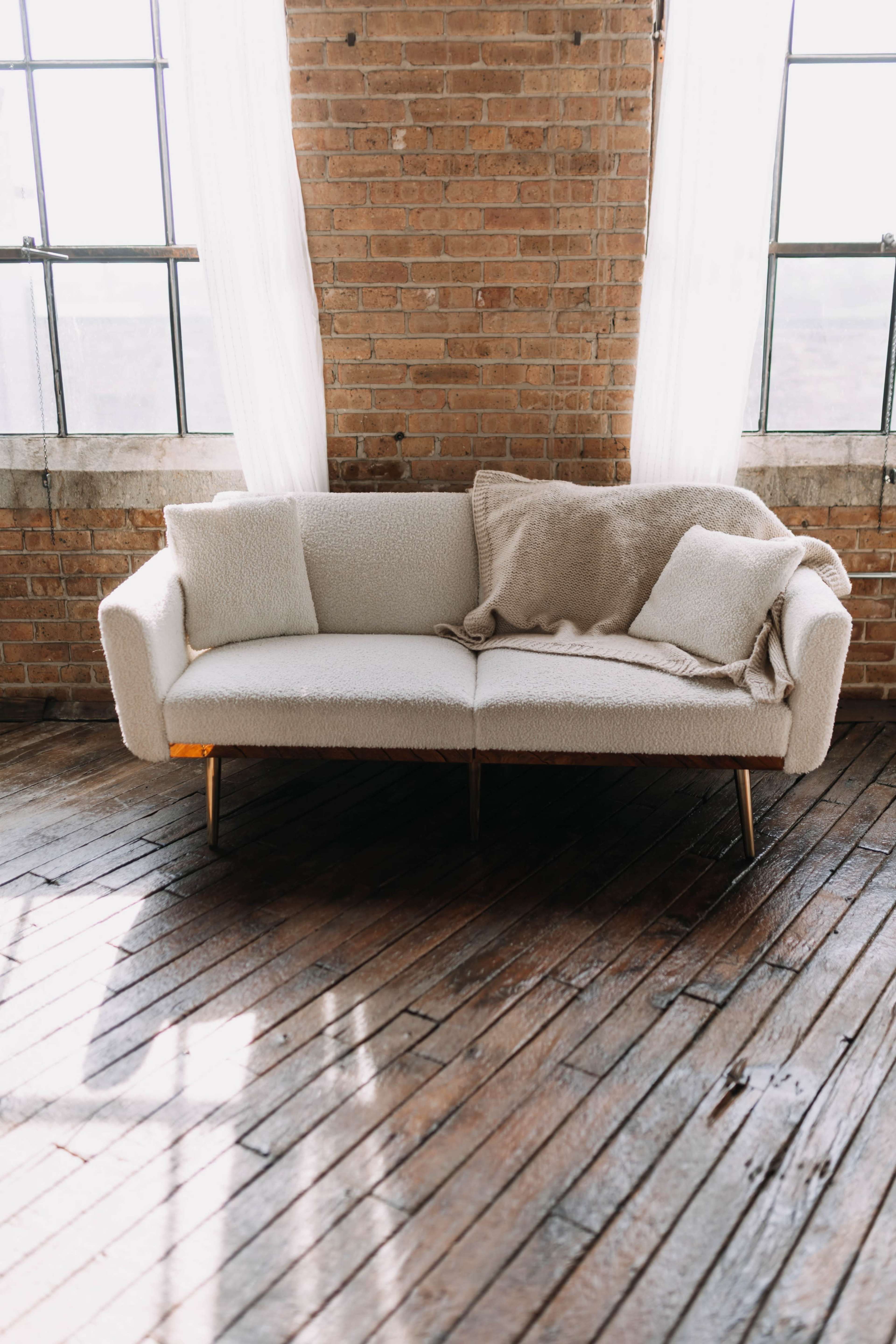 A light-colored couch with a textured fabric and throw pillows is positioned on a wooden floor, illuminated by natural light streaming through large windows.