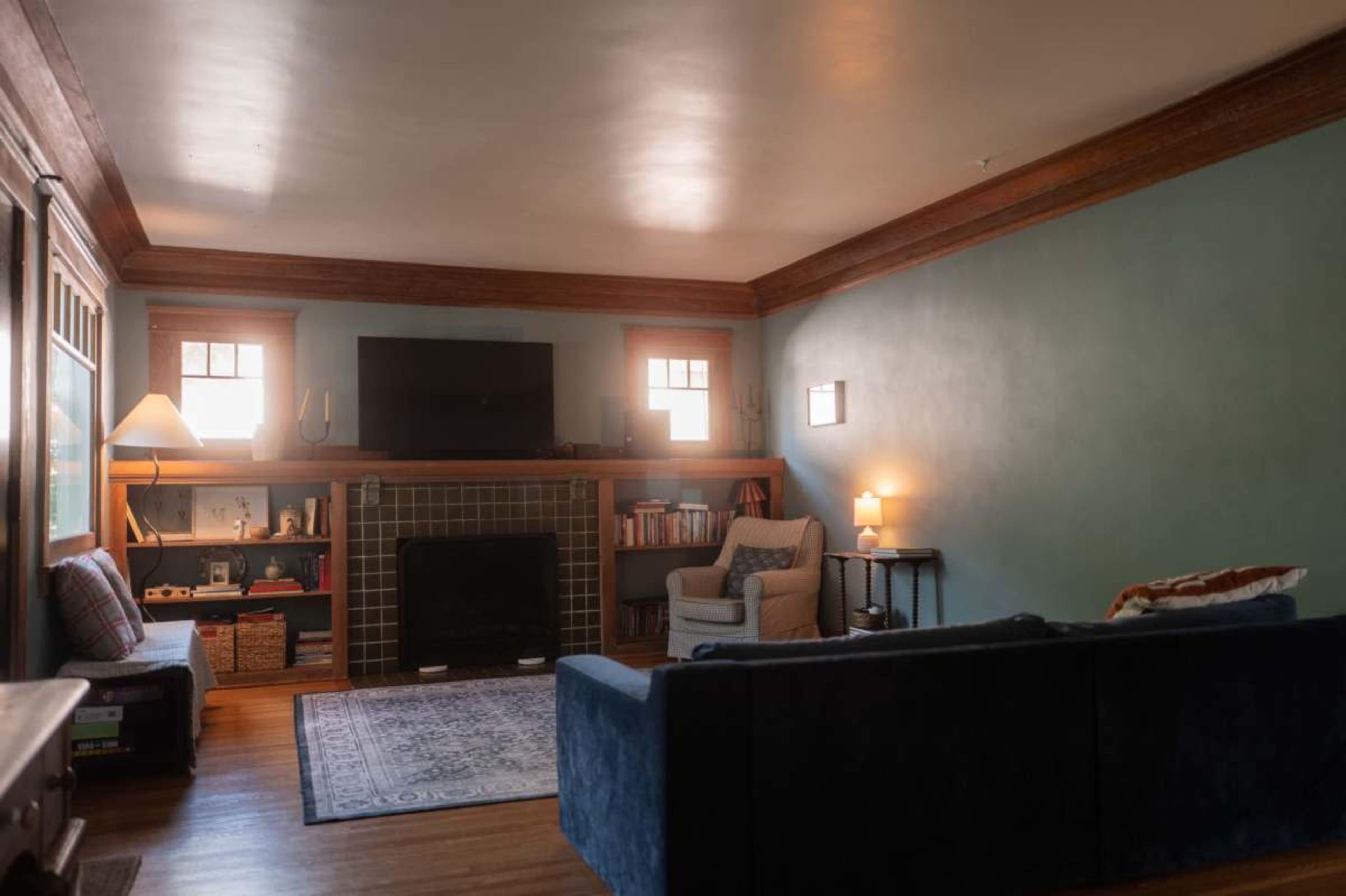 The image shows a cozy living room with a blue sofa, a fireplace, and a television mounted on the wall, surrounded by bookshelves and a floor lamp.