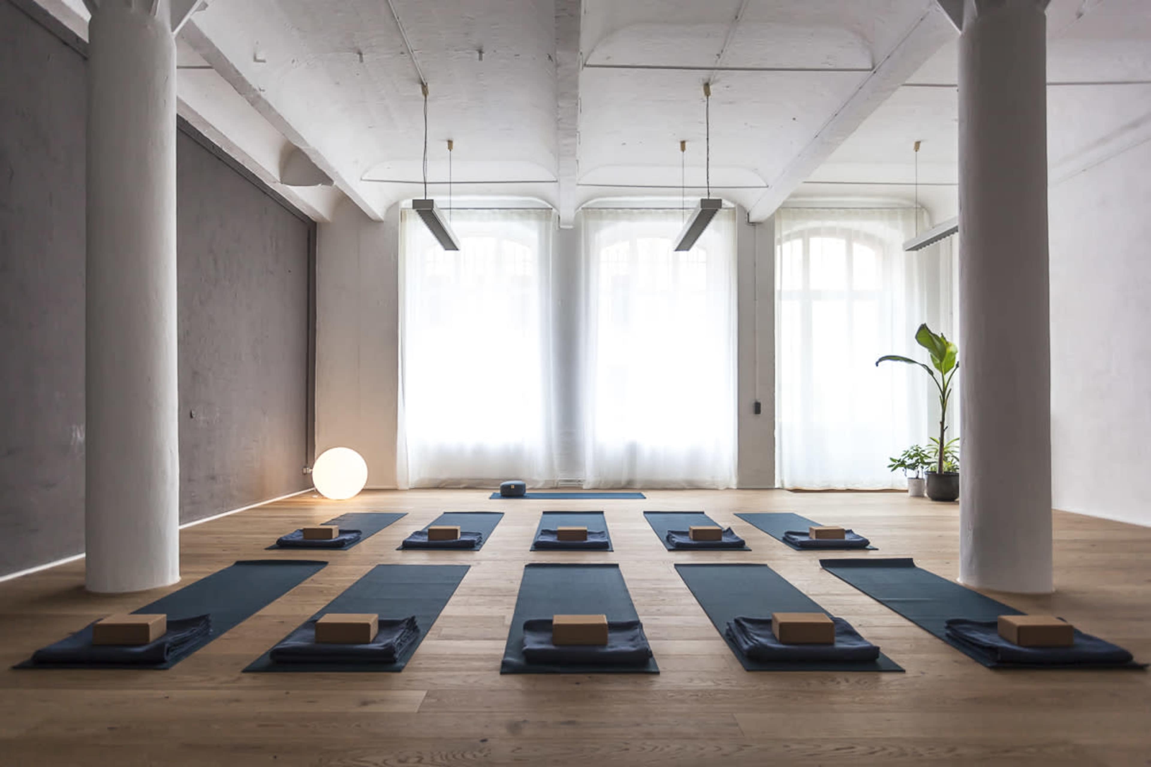 The image shows a yoga studio with neatly arranged mats and blocks on a wooden floor, illuminated by natural light filtering through large windows.