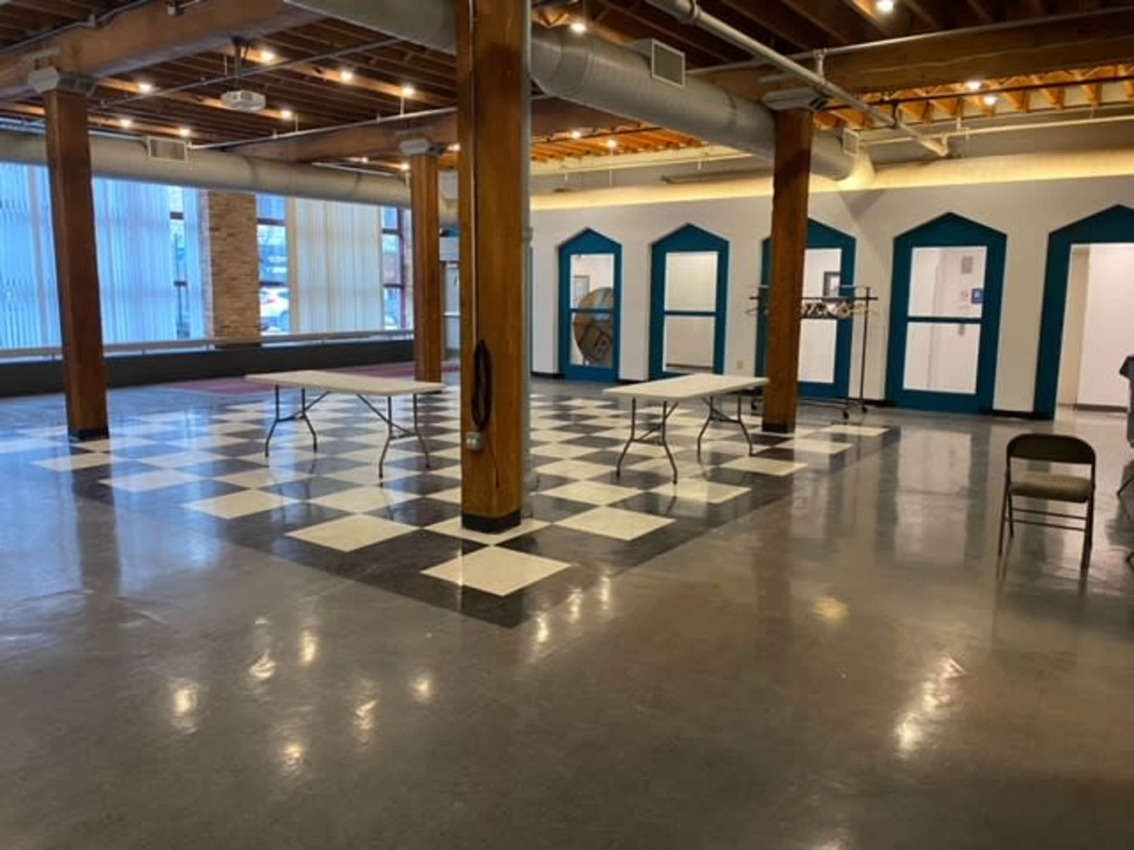 An empty room with a checkered floor, several tables set up, and large windows allowing natural light.