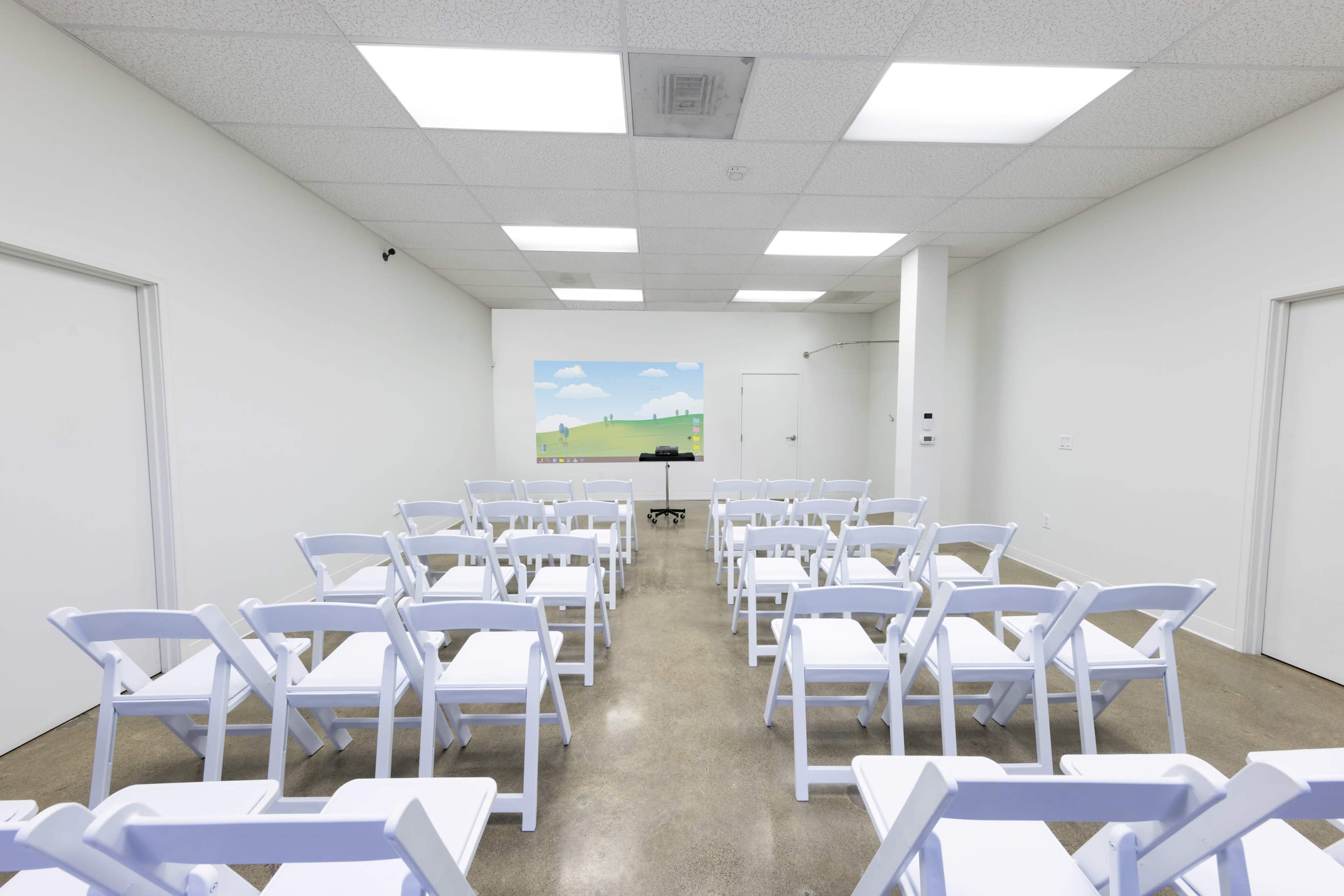 A sparsely furnished room features white folding chairs arranged in rows facing a wall-mounted screen displaying a simple landscape scene.