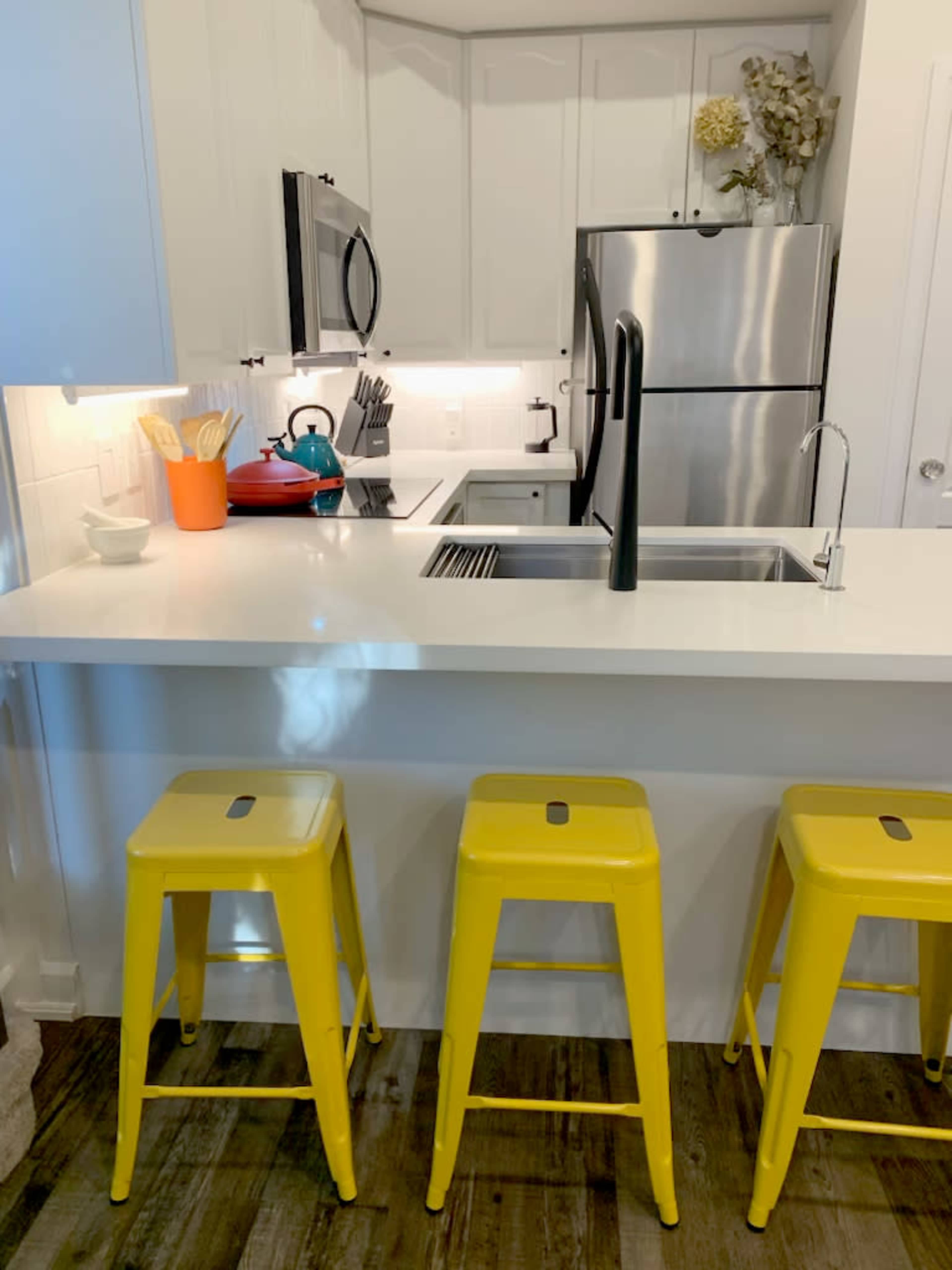 The image shows a modern kitchen with a white countertop, stainless steel appliances, and three yellow bar stools at a small seating area.