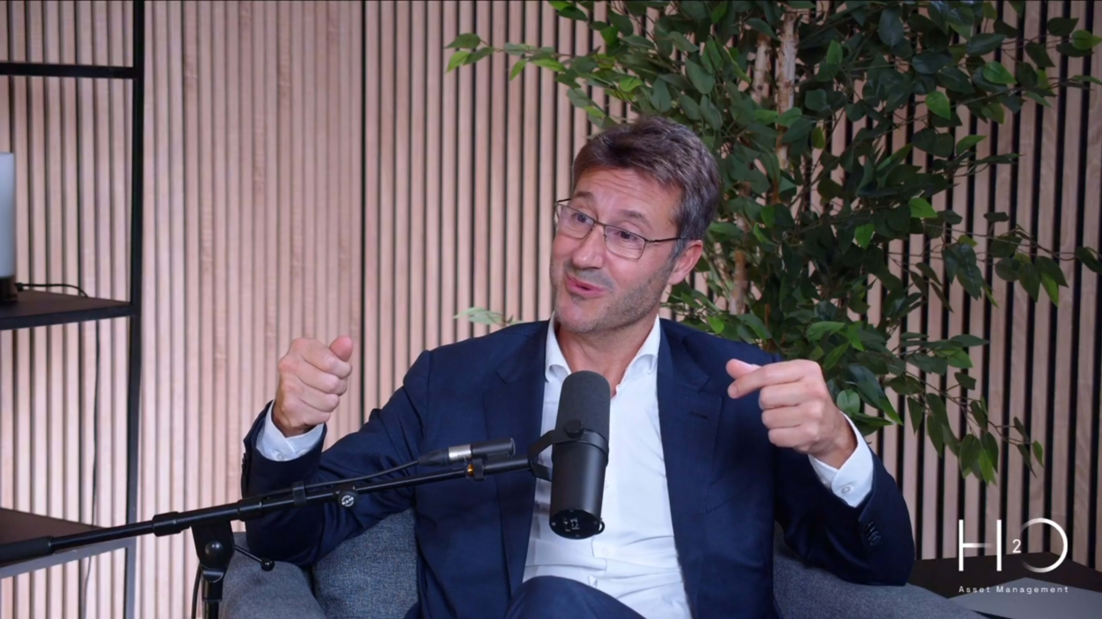 A man in a blue suit gestures while seated in a modern setting with plants and a microphone in the foreground.