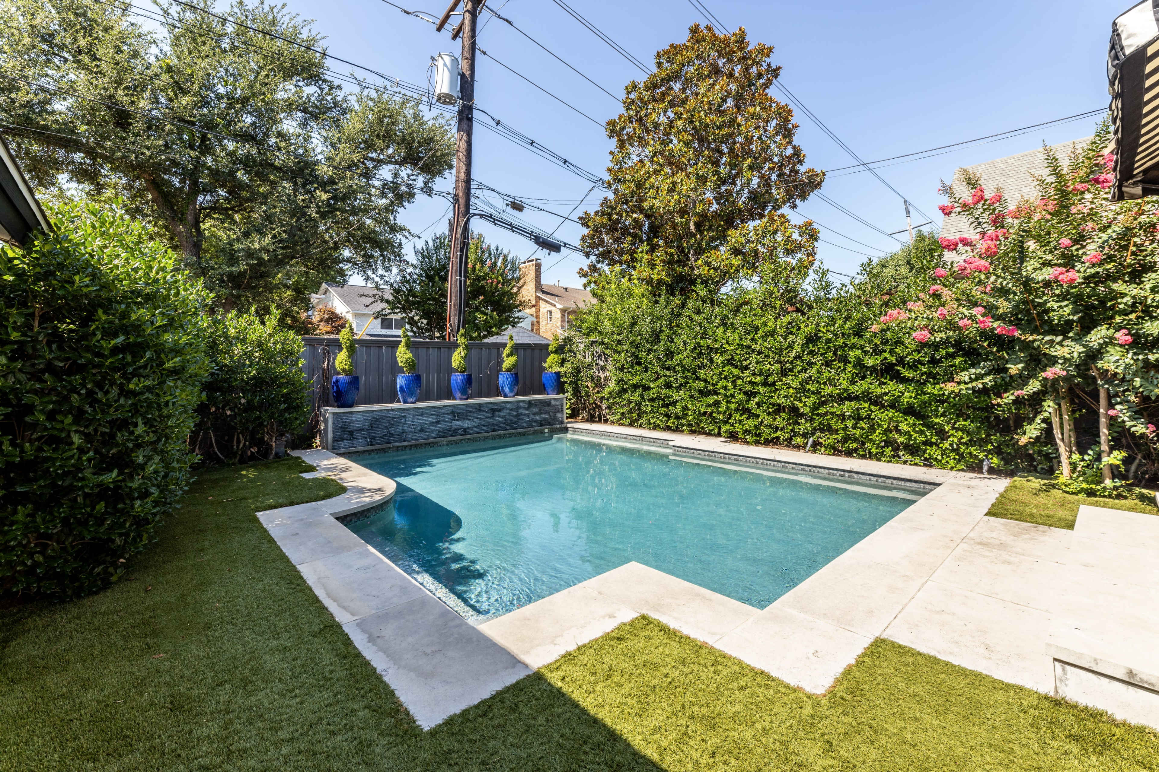 The image shows a rectangular swimming pool surrounded by green grass and tall hedges, with a background of flowering plants and utility poles.