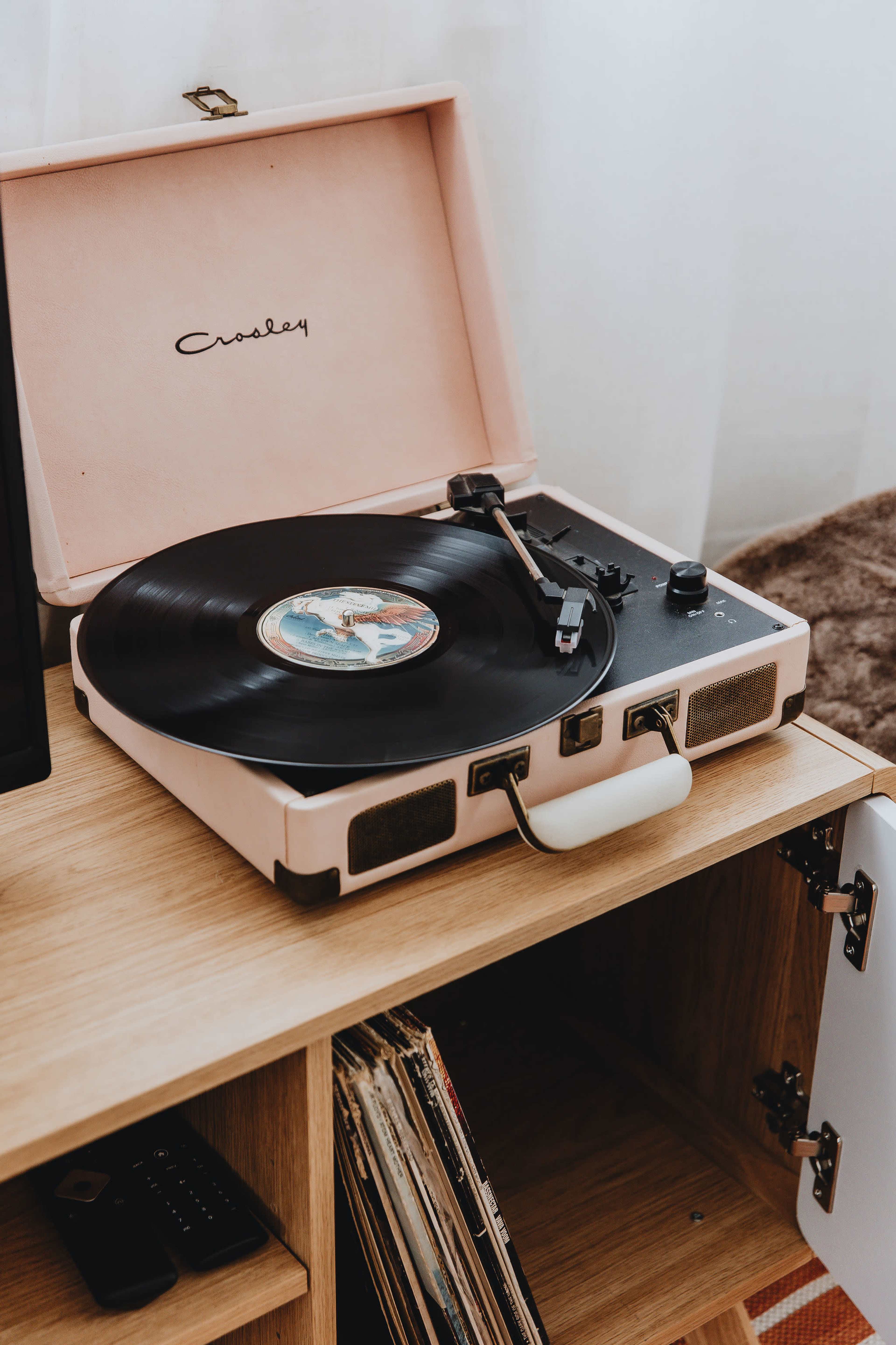 A pink portable turntable with a black record is placed on a wooden shelf next to a stack of vinyl records.