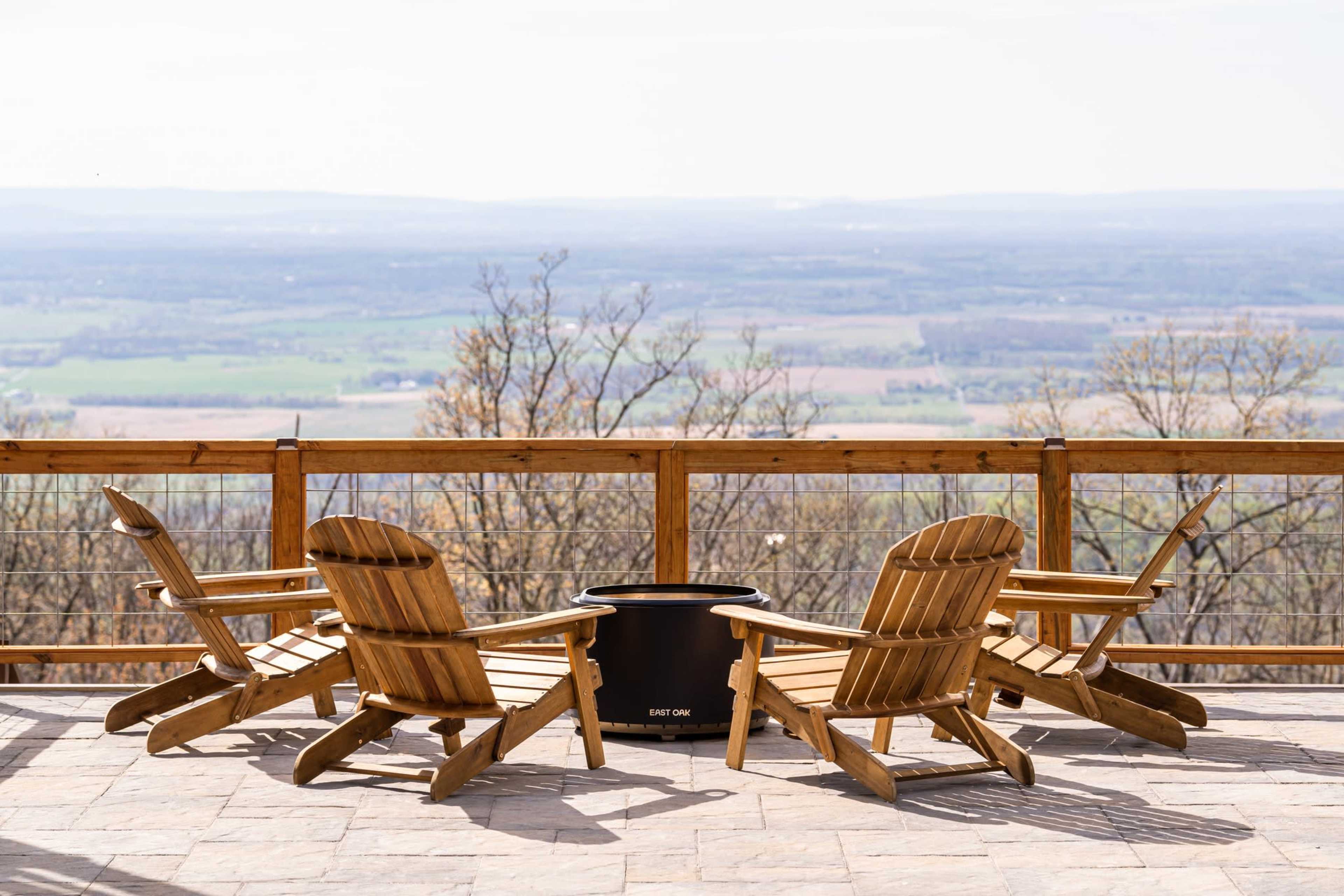Four wooden Adirondack chairs are arranged in a circle around a black fire pit on a stone patio overlooking a scenic valley.