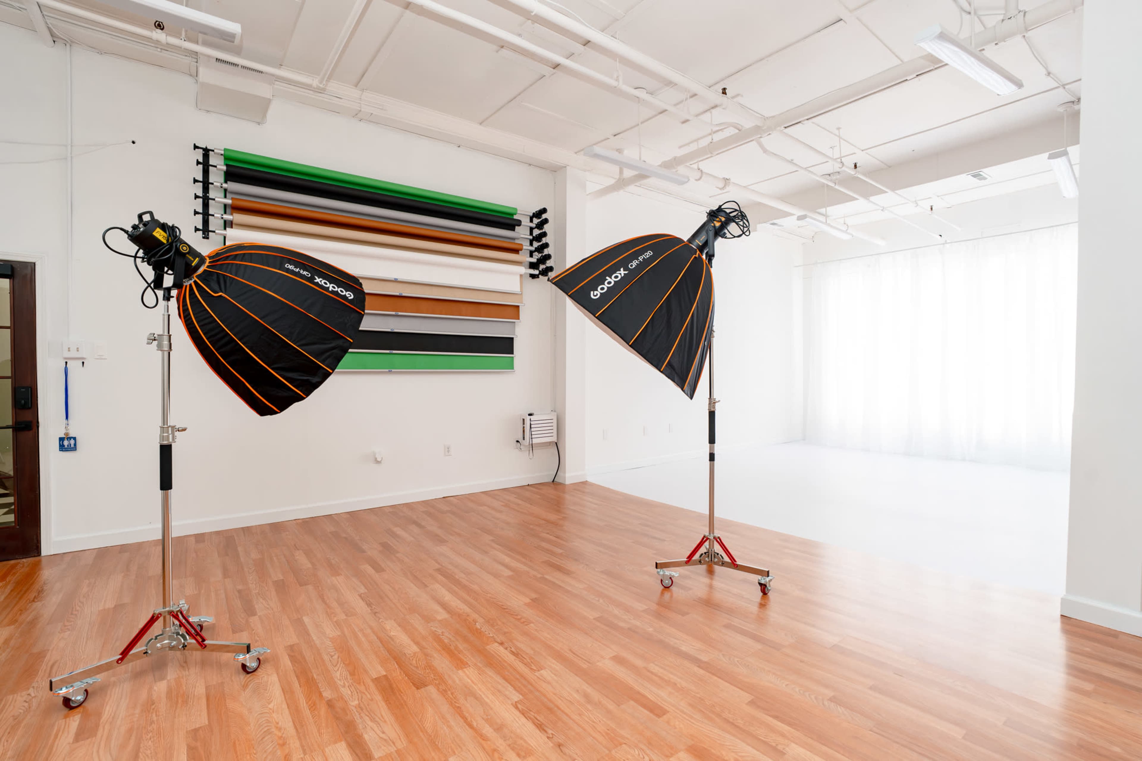 The image shows a spacious studio with wooden flooring, featuring two softbox light setups and a wall with various colored backdrops.