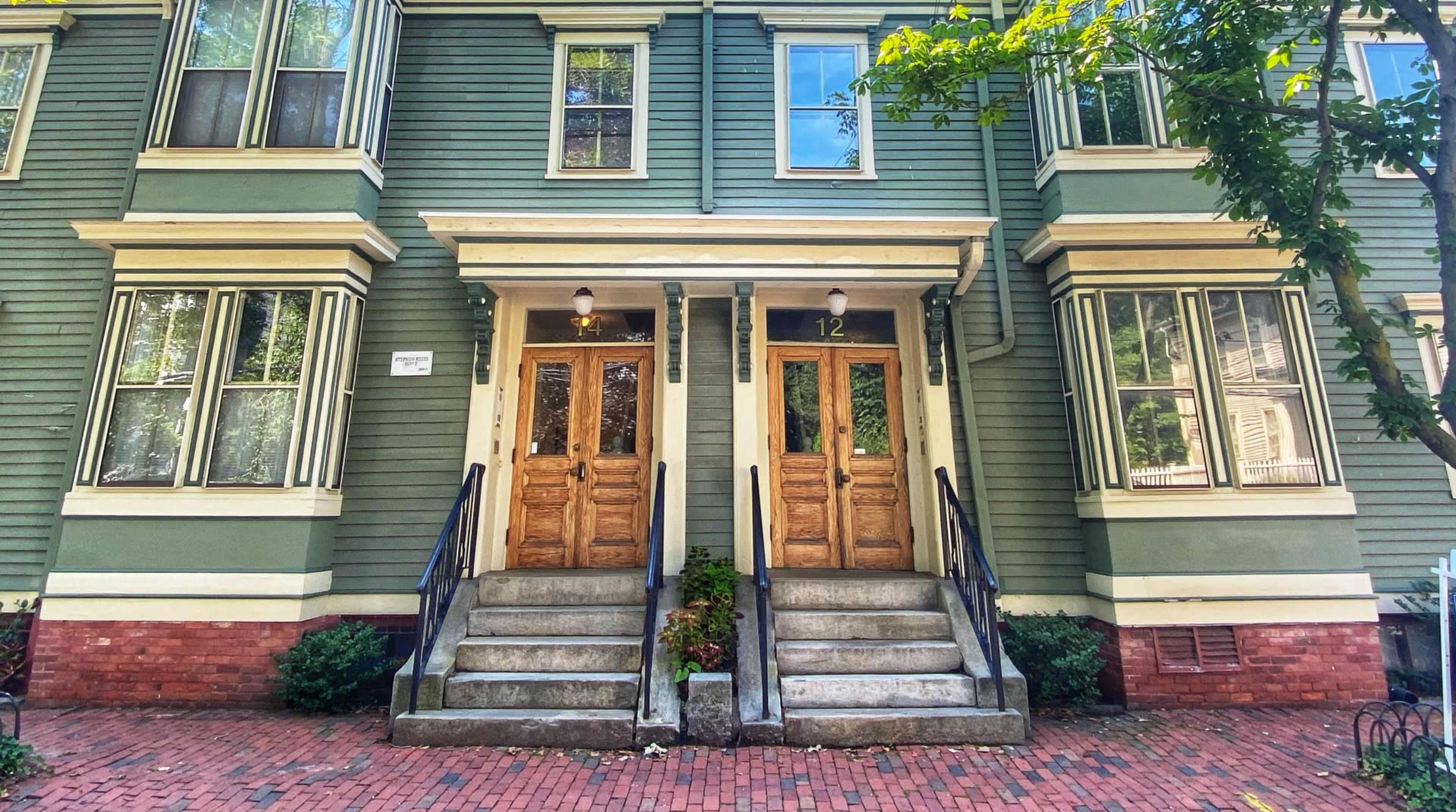 A pair of wooden doors at the entrance of a green multi-story building, flanked by a staircase on either side and surrounded by brick paving and small shrubs.