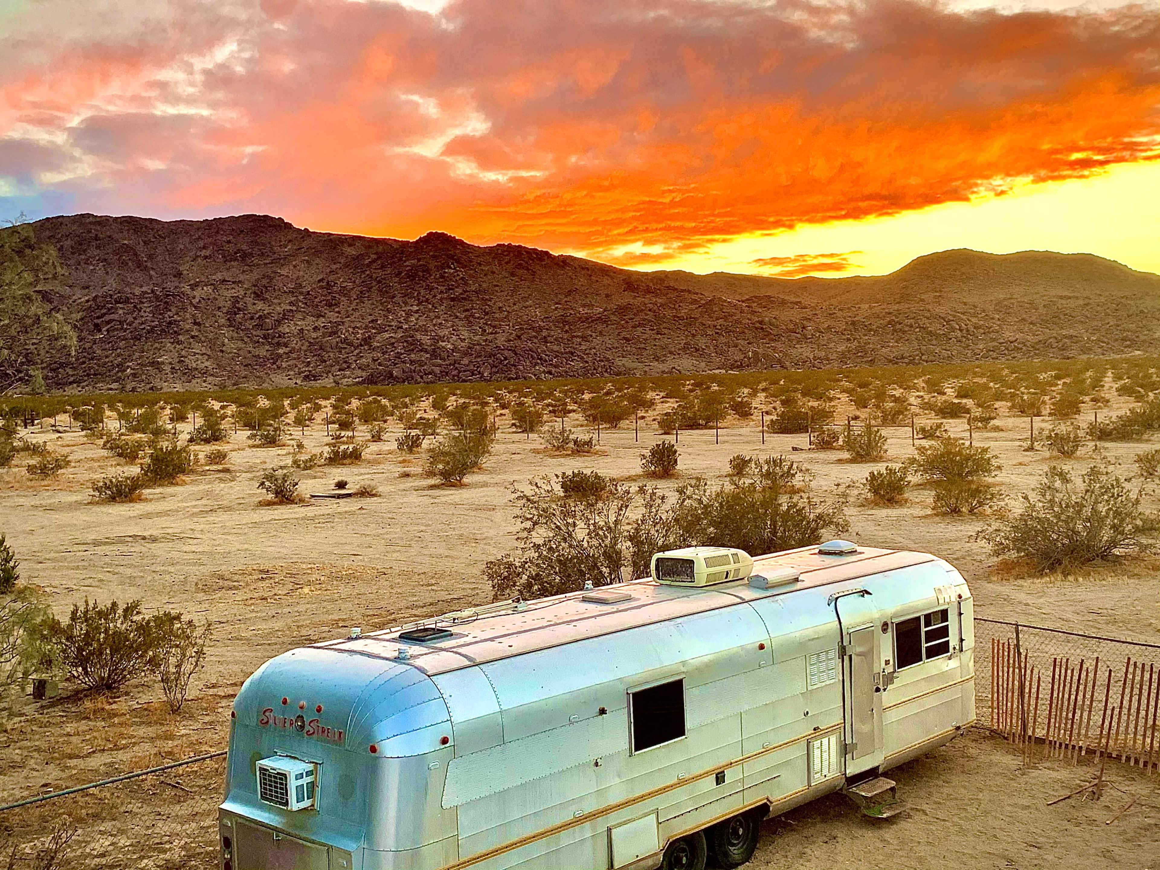 An Airstream trailer is parked in a desert landscape with a vibrant sunset illuminating the sky.