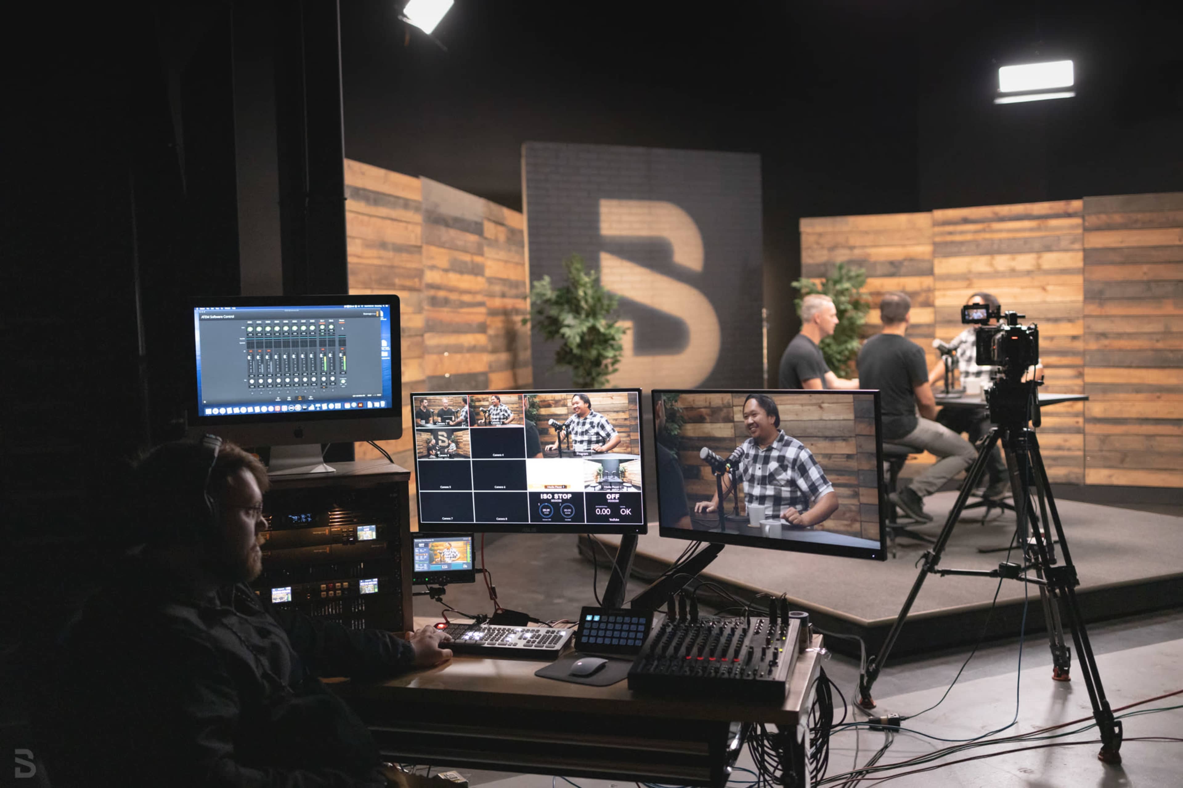 A video production setup shows a person operating equipment in front of multiple monitors while a podcast is being filmed in a studio with wood-paneled walls.