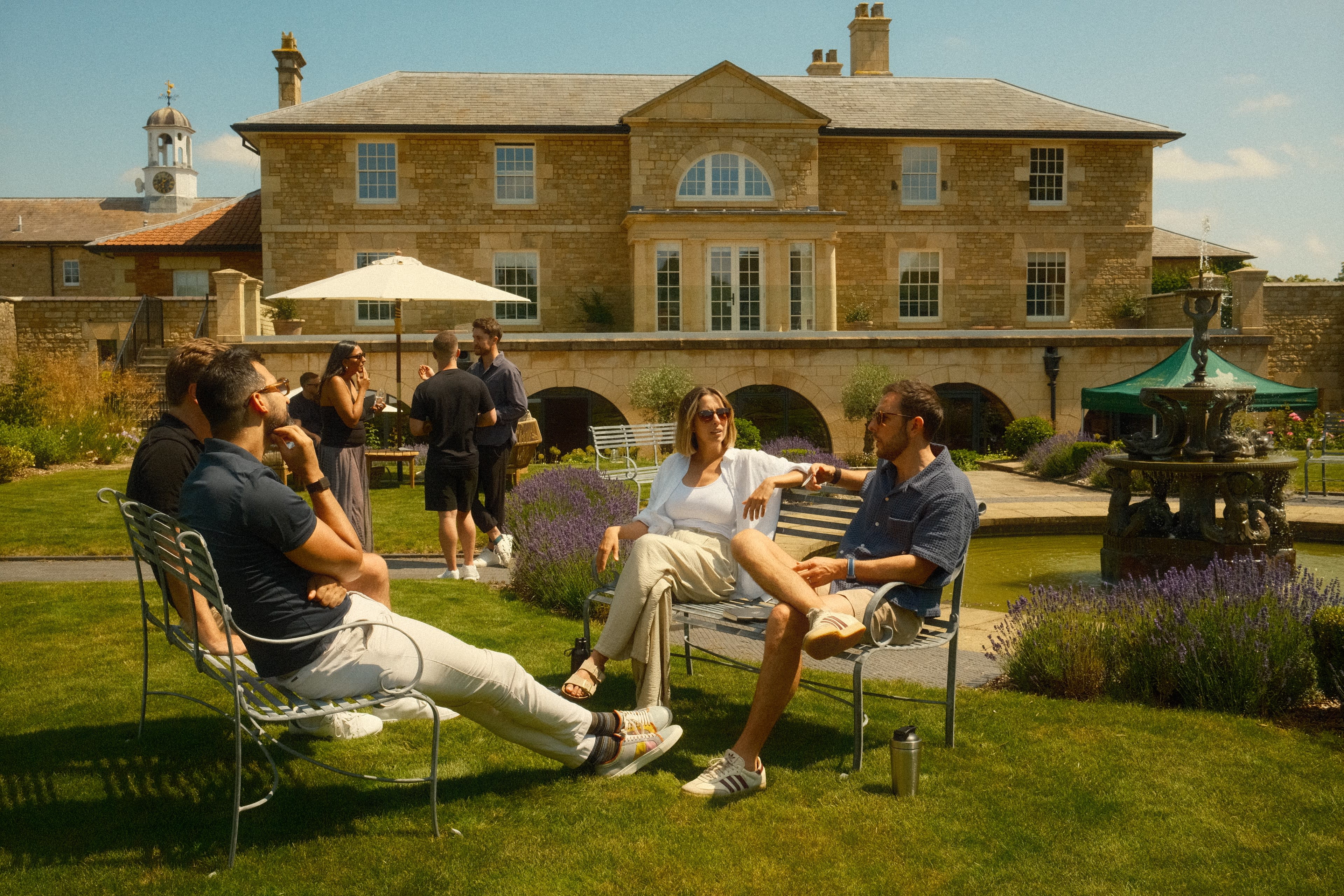 A group of people is gathered in a garden, seated on chairs in front of a large, two-story stone building with a fountain and landscaping.