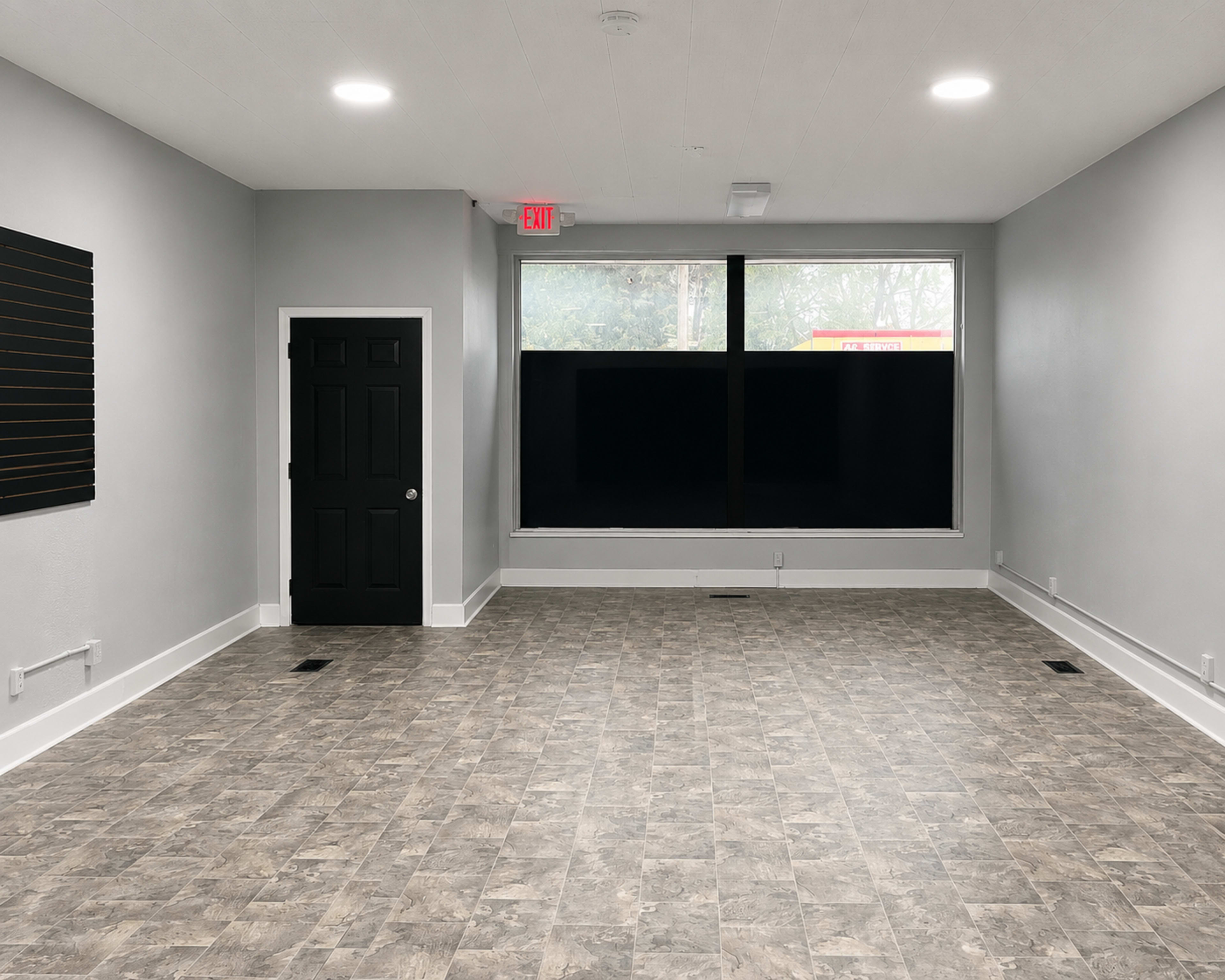 The image depicts an empty room with gray walls and tiled flooring, featuring a black door and a large window with a dark covering.