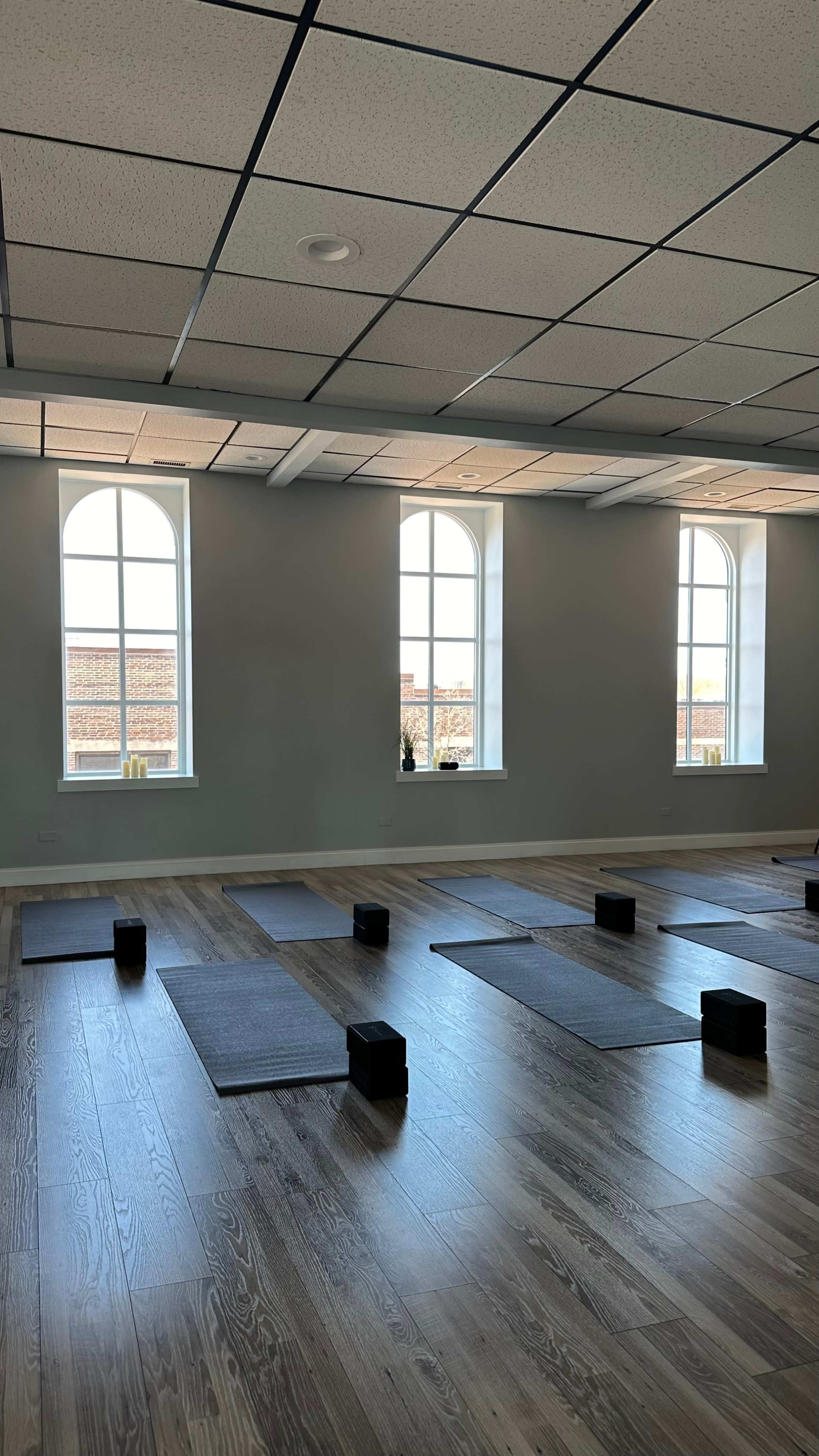 A yoga studio with several mats arranged on the wooden floor beneath large, arched windows.