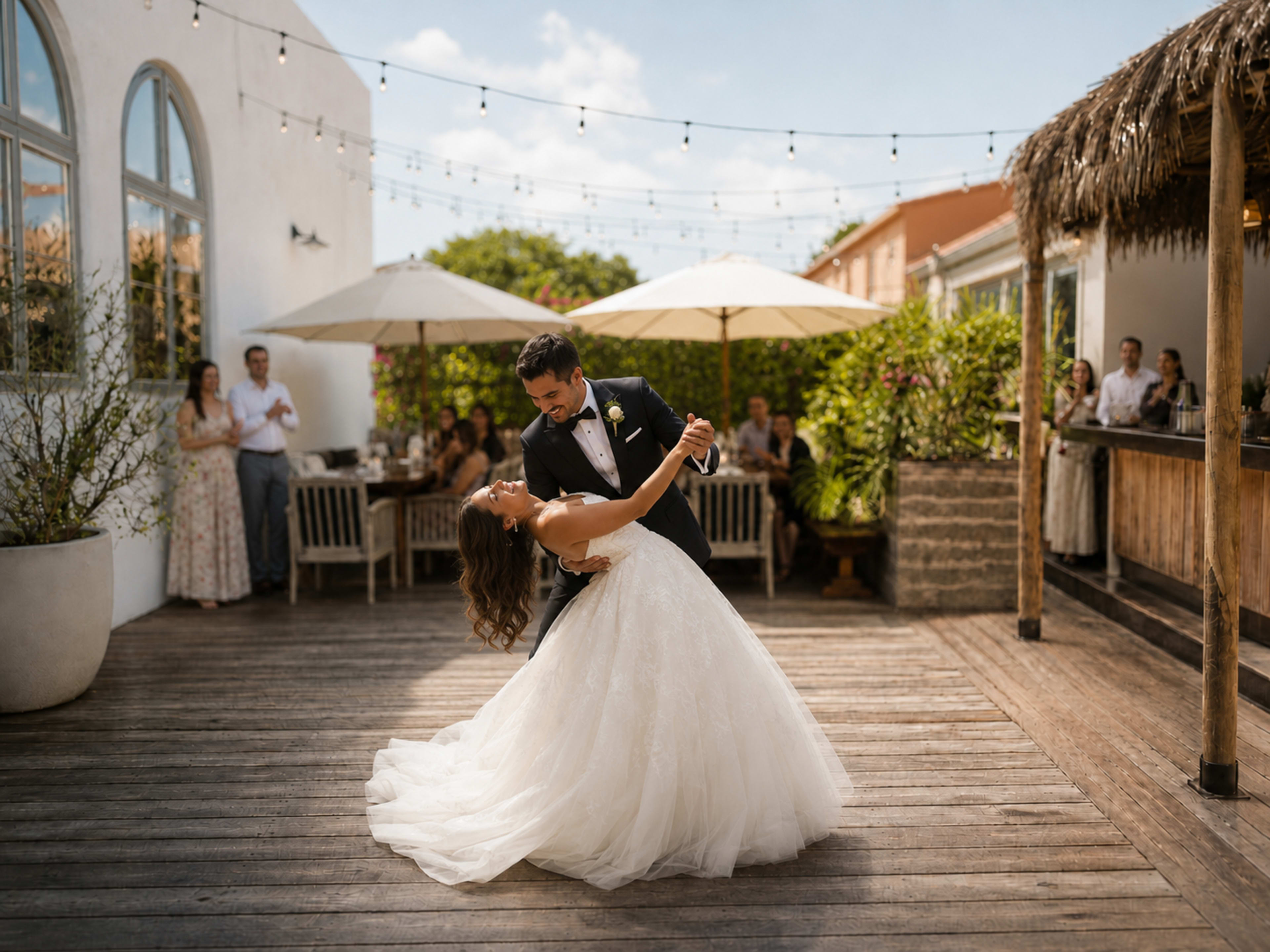 A bride in a white wedding gown and a groom in a black tuxedo are dancing together in an outdoor setting with guests seated at tables in the background.
