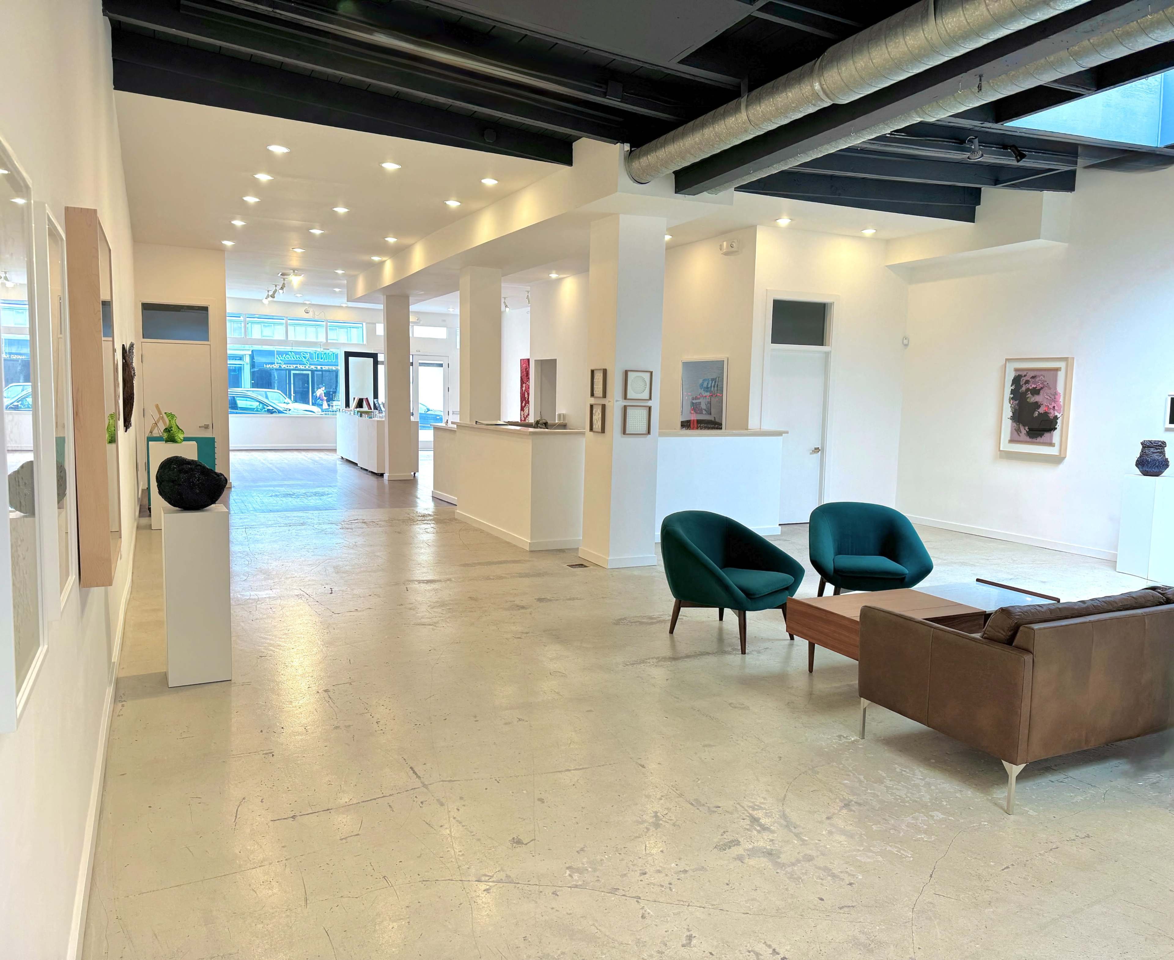 The image shows a modern art gallery with white walls, polished concrete floors, and several artworks displayed throughout the space, including seating areas.