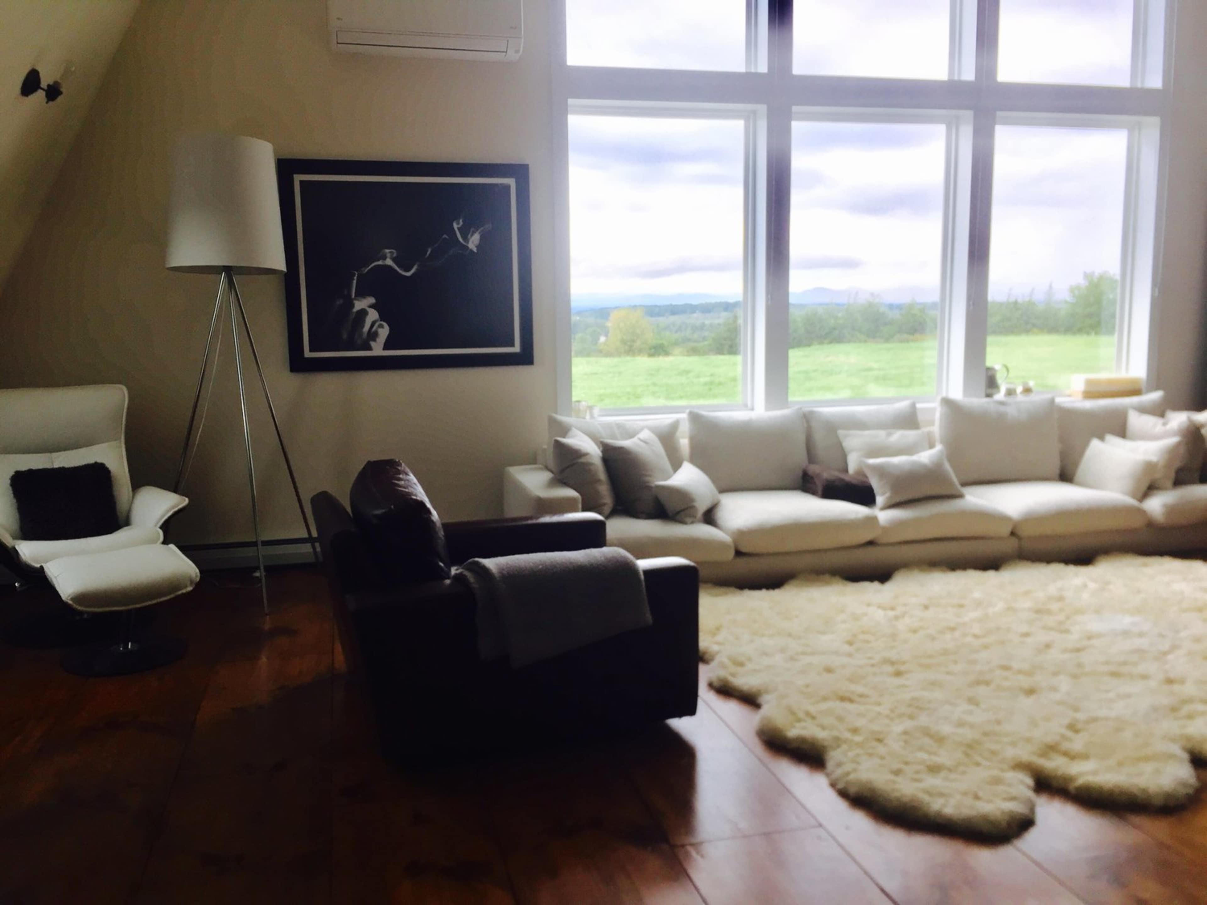 A modern living room with a large sectional sofa, a brown leather armchair, a white floor lamp, and large windows offering a view of a green landscape.