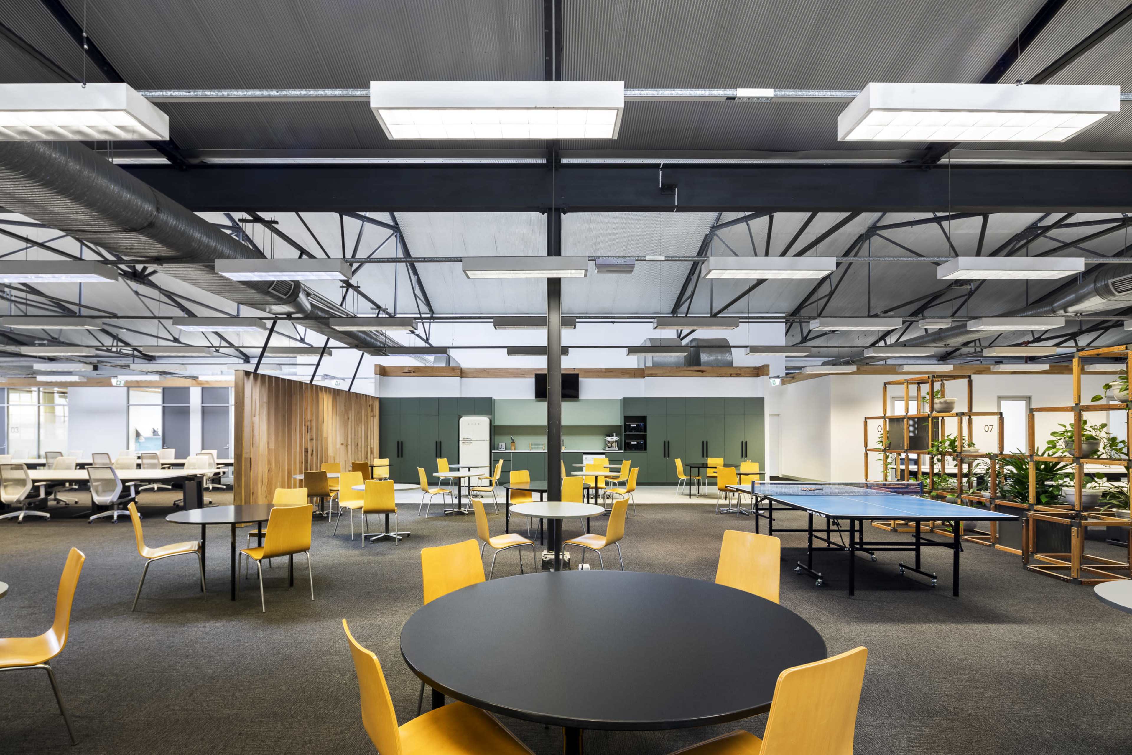 The image shows a modern office space with yellow chairs, black round tables, and a ping pong table, featuring exposed ceilings and a green accent wall.