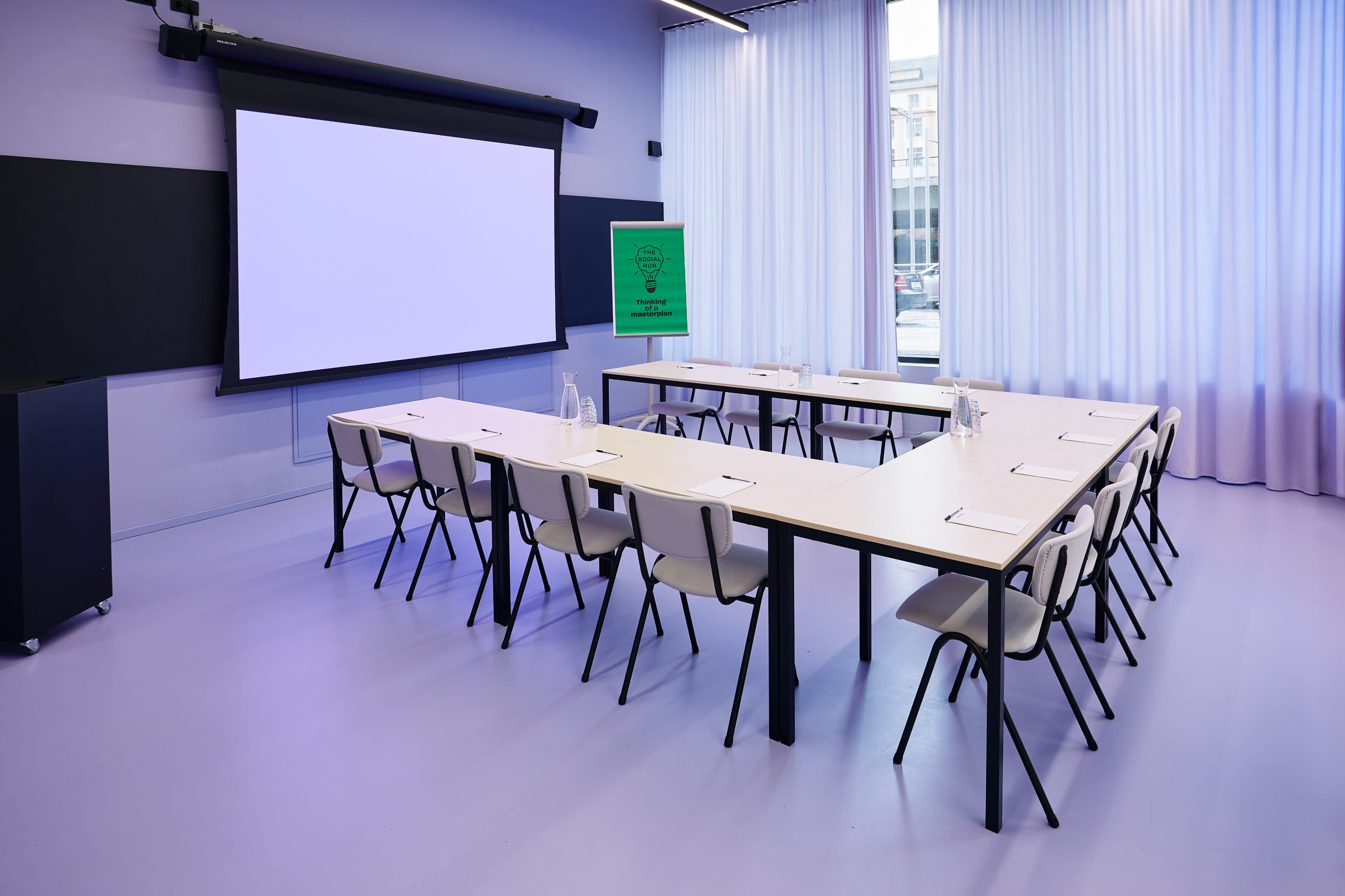 A modern conference room features a rectangular table surrounded by chairs, with a projector screen at the front and large windows allowing natural light.