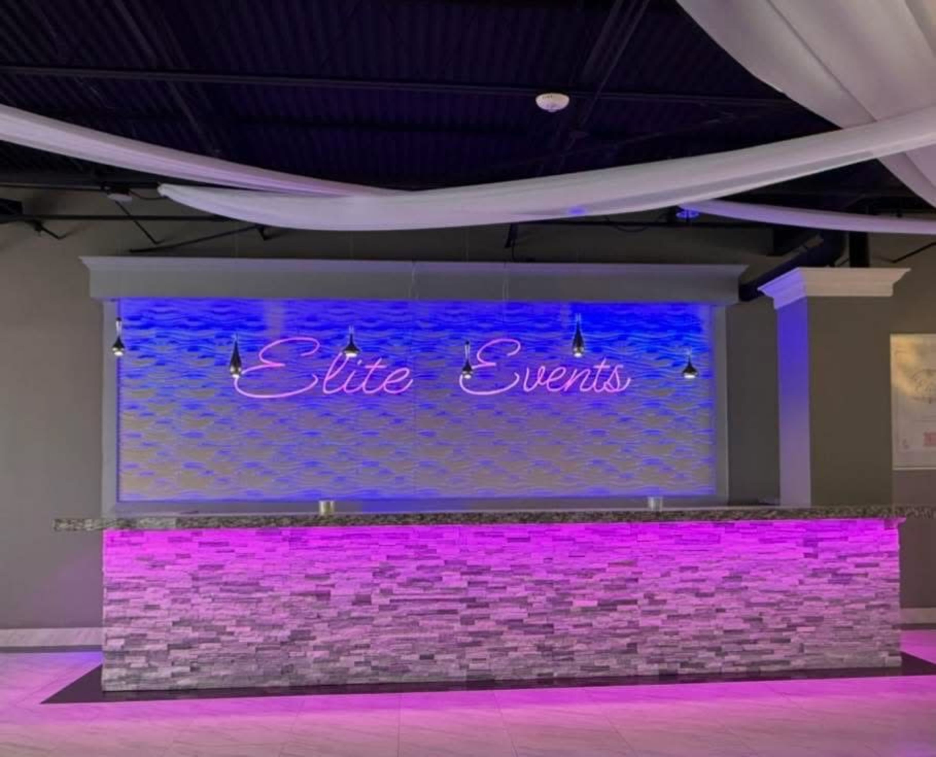 A modern event space features a granite-topped bar with a neon sign reading "Elite Events" and ambient lighting in shades of blue and pink.
