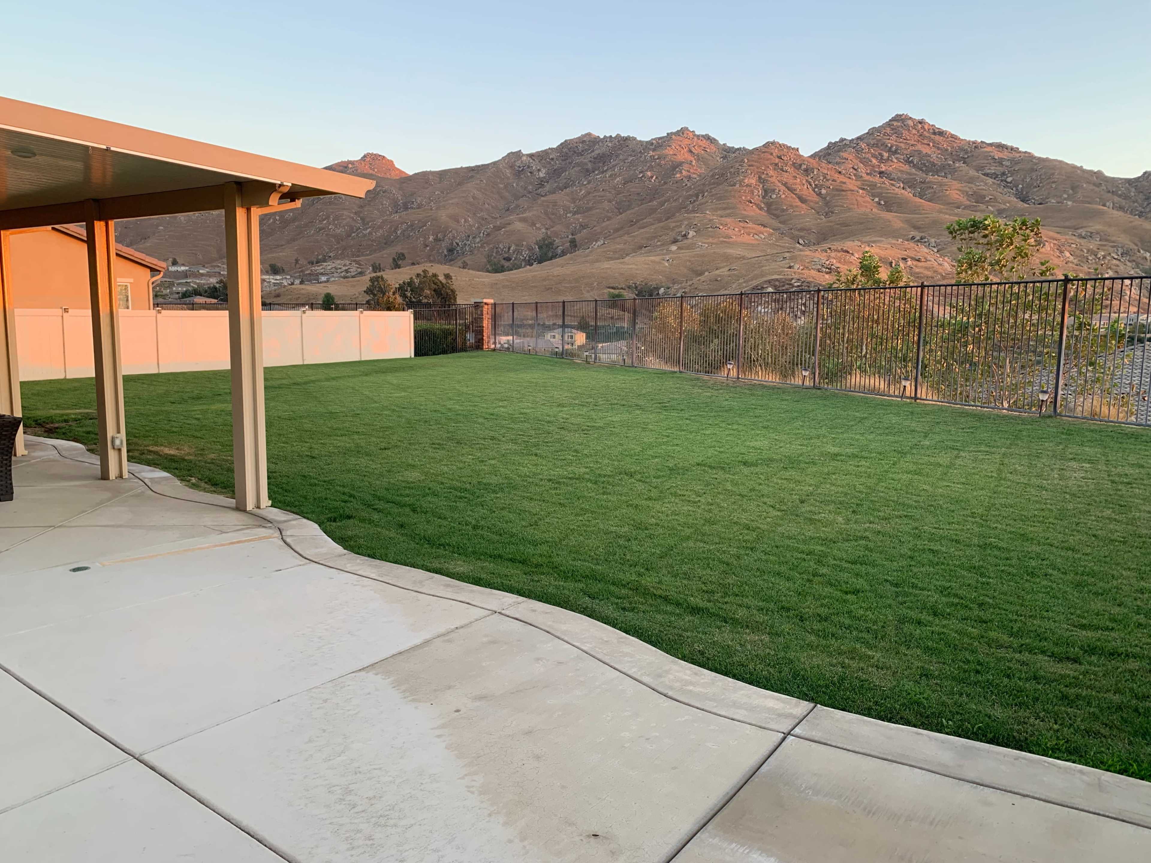 The image shows a backyard with a neatly maintained green lawn, a concrete patio, and a view of mountains in the background.
