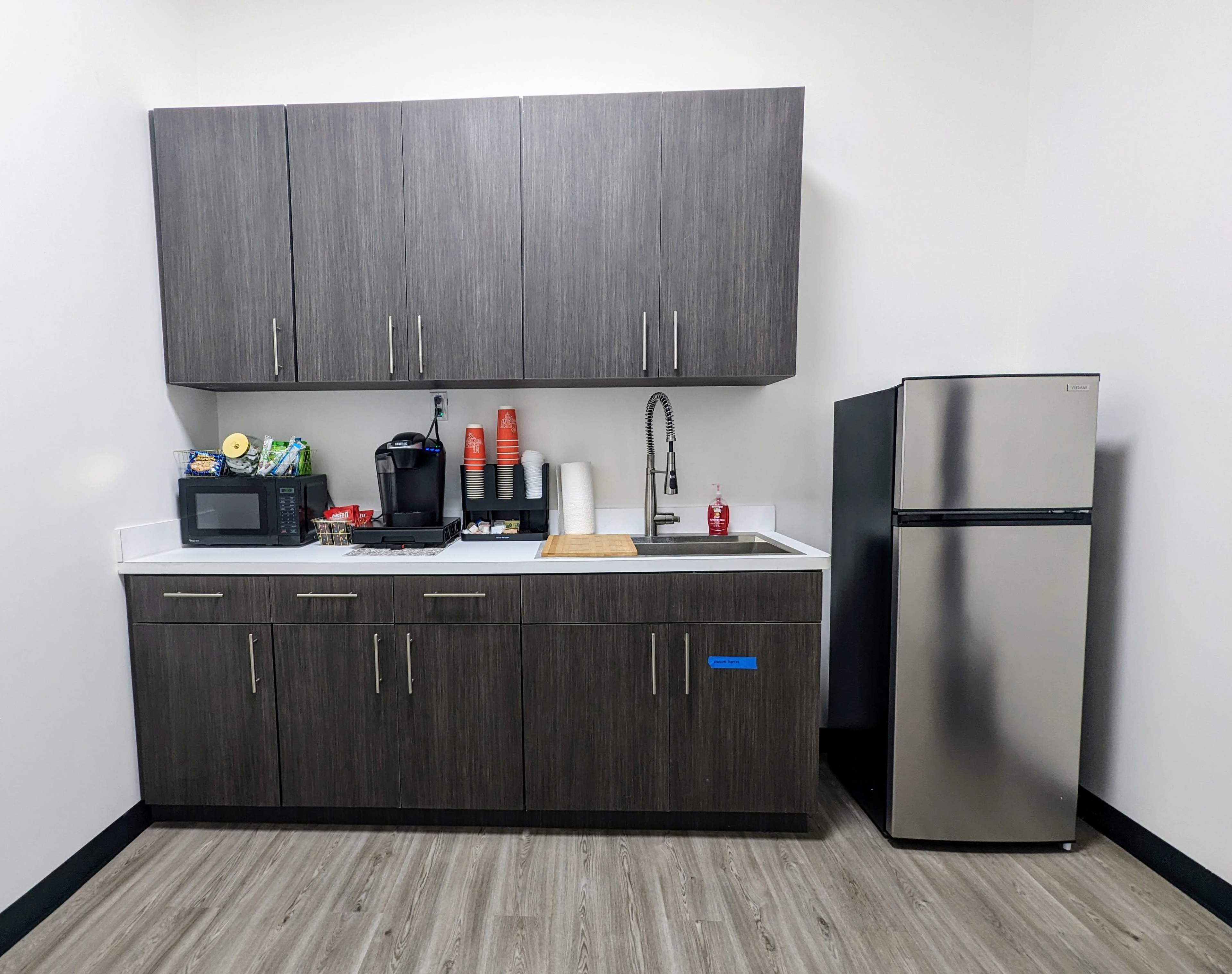The image shows a modern kitchen corner with dark cabinetry, a microwave, a coffee maker, and a stainless steel refrigerator.