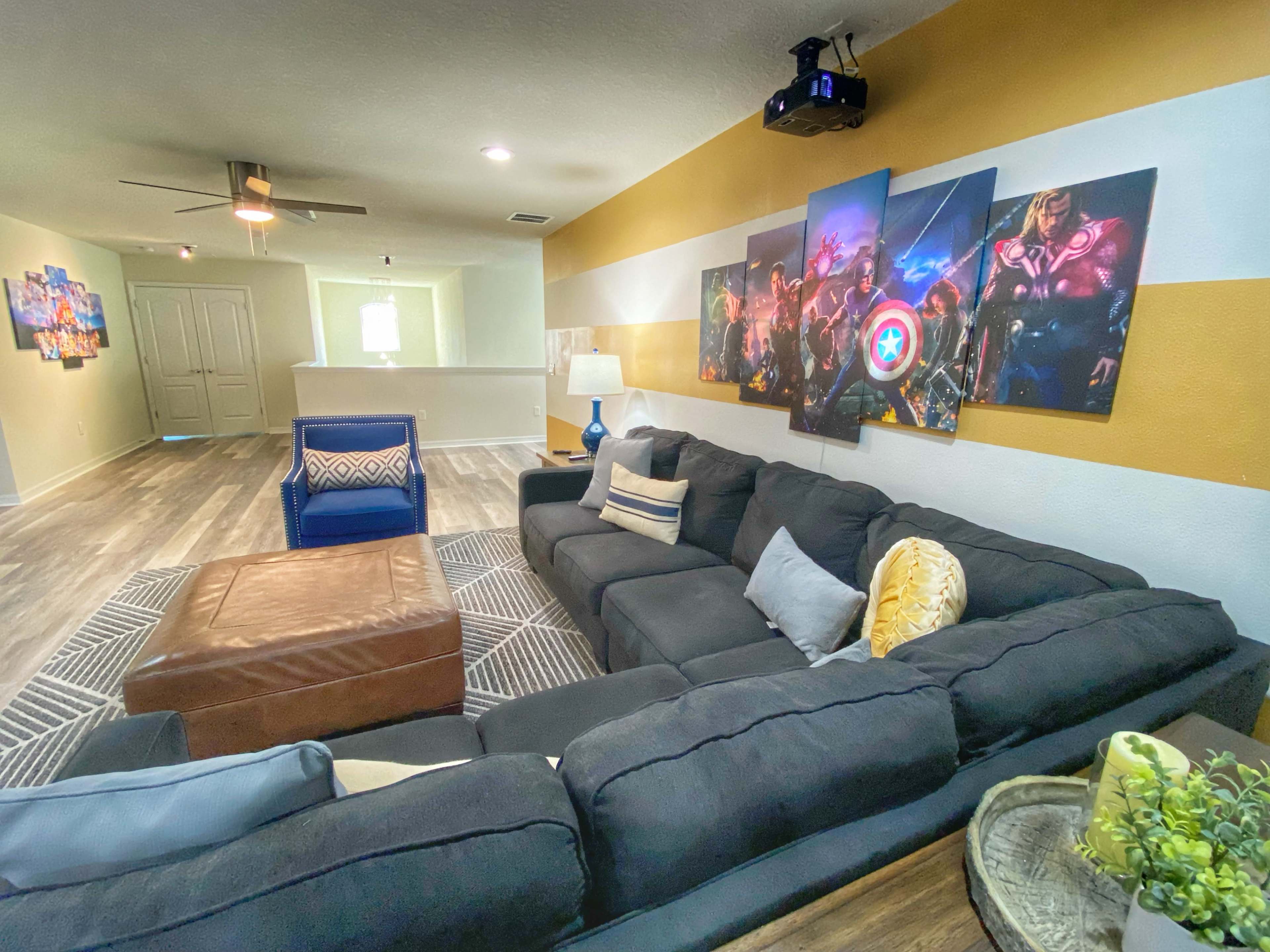 A modern living room features a large sectional couch, a coffee table, and wall art depicting superheroes, with a projector mounted on the ceiling.