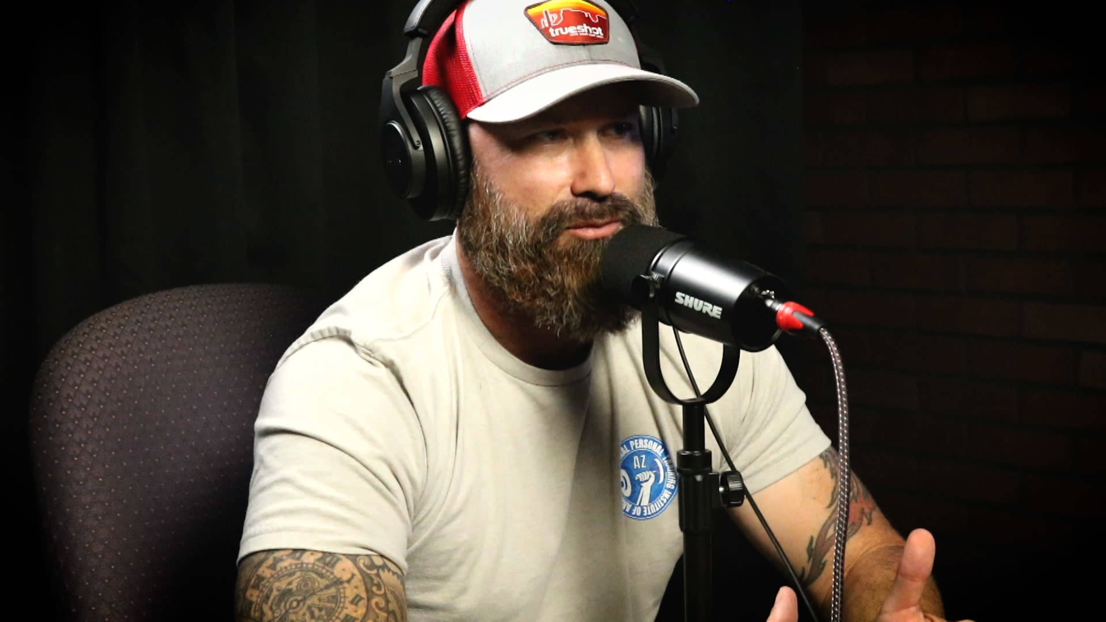 A man with a beard and tattoos speaks into a microphone while wearing headphones and a cap.