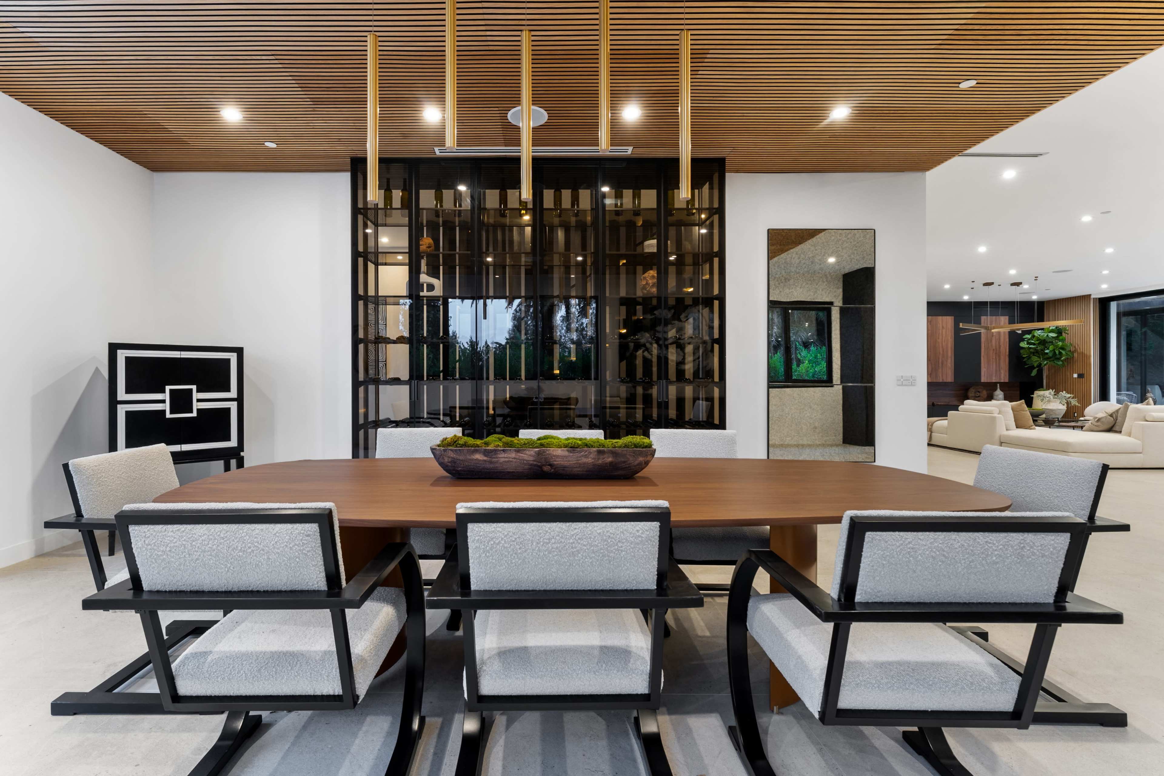 A modern dining room features a large wooden table surrounded by six upholstered chairs, with a backdrop of stylish glass and wood paneling.