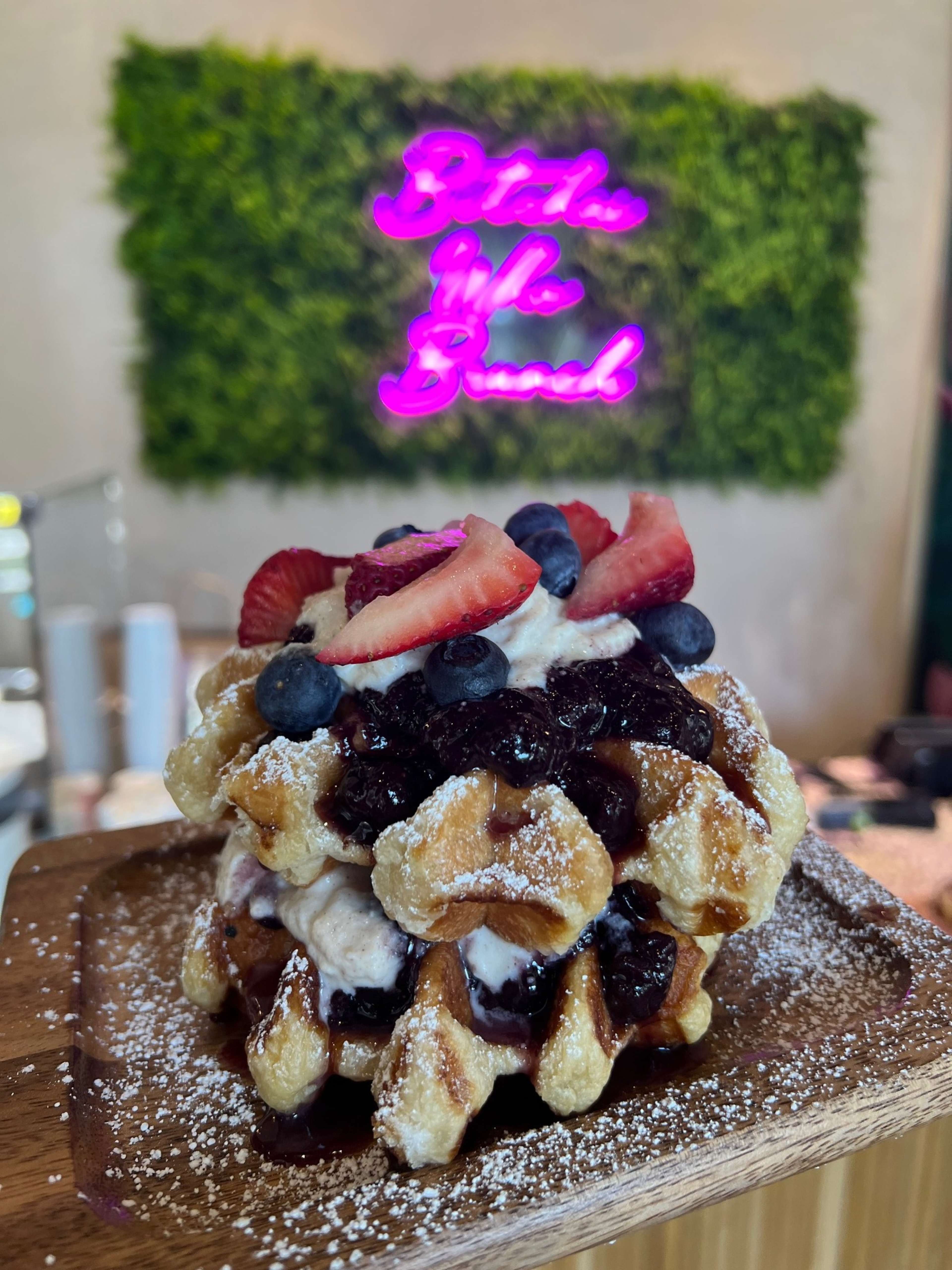 A stack of waffles topped with whipped cream, mixed berries, and a drizzle of berry sauce is presented on a wooden plate against a backdrop of a neon sign.