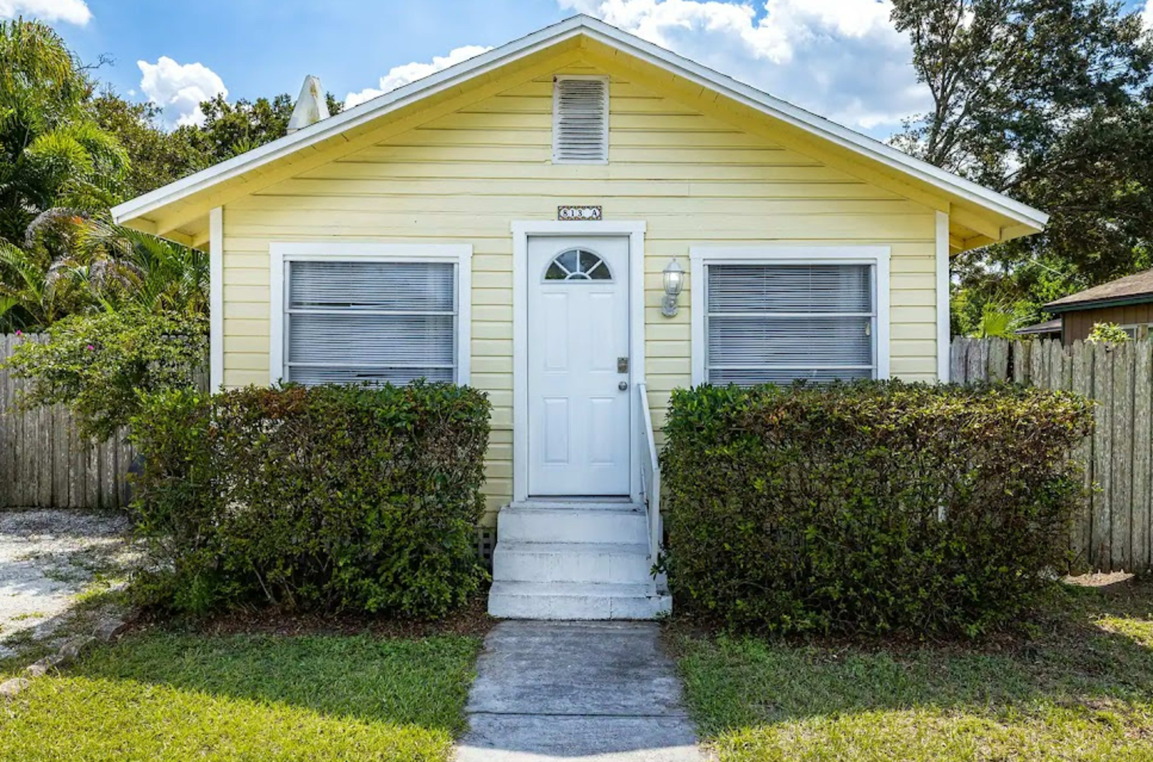 A small, yellow house with a white front door and shrubs on either side is set against a blue sky.