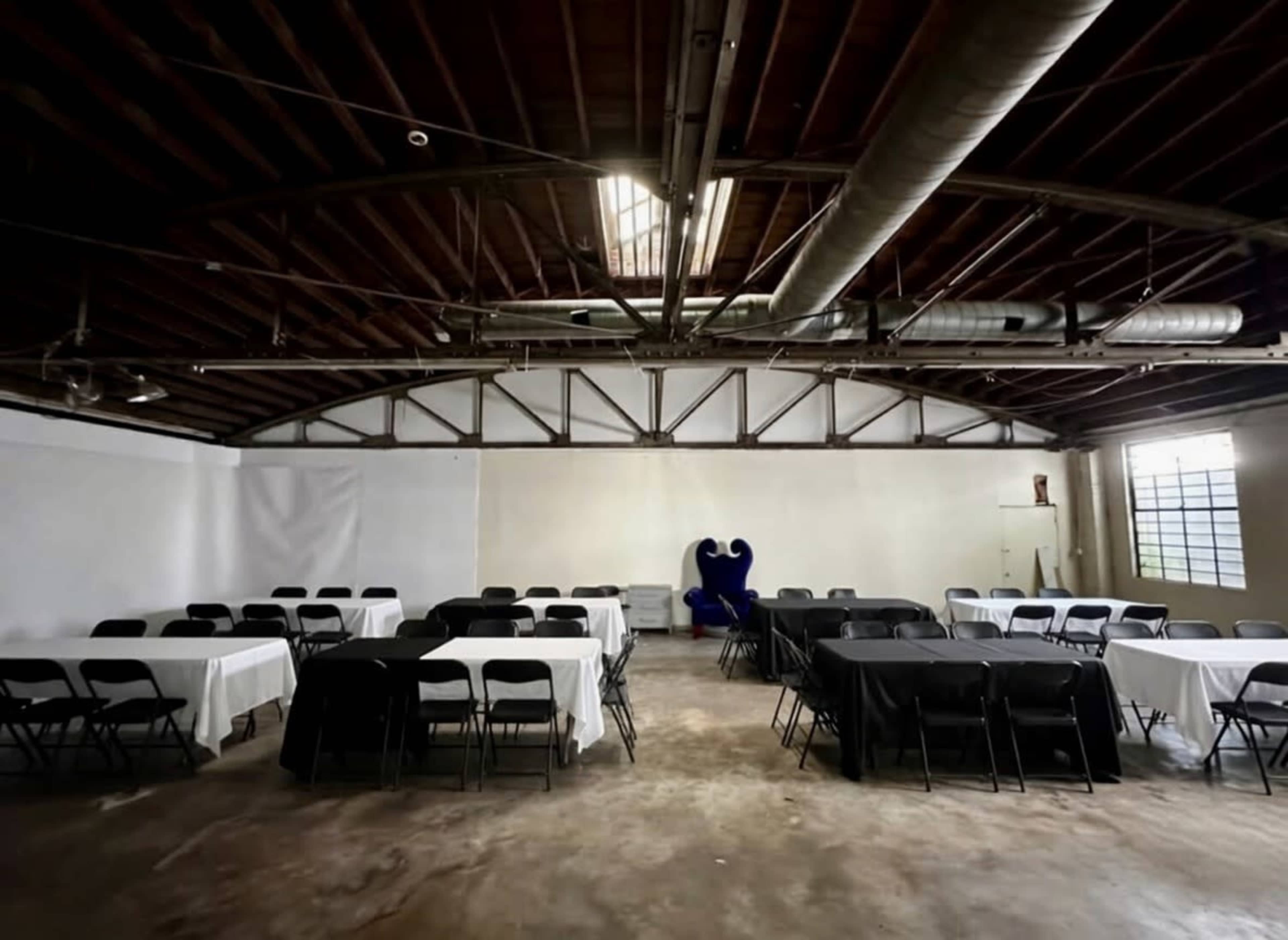 The image shows an empty event space with rectangular tables covered in black and white tablecloths arranged in a symmetrical layout and a large blue chair in the background.