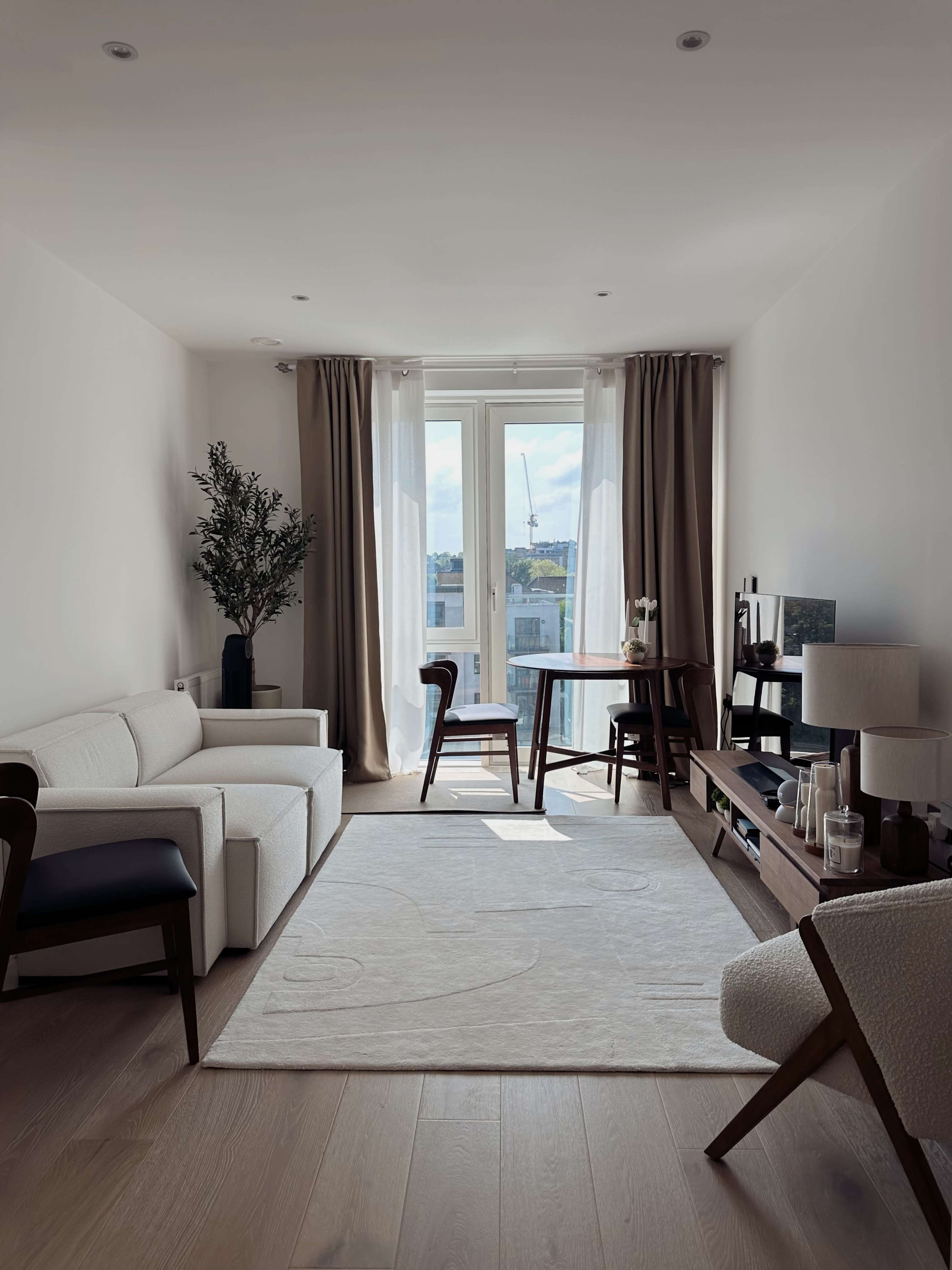 A well-lit living room features a white sofa, a wooden table with chairs, and a large window with light curtains, overlooking an outdoor view.
