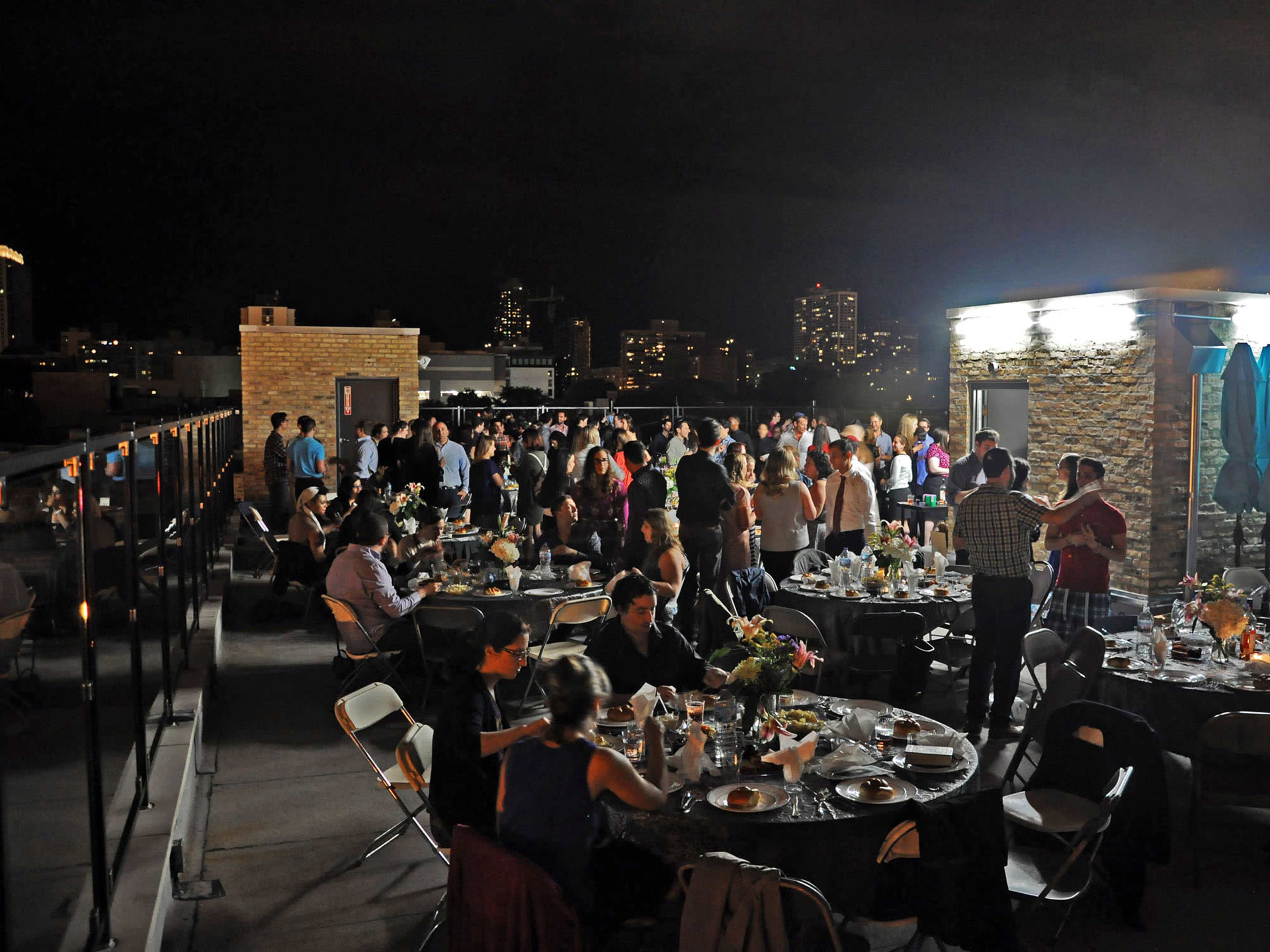 A large group of people socializes at a rooftop event, with tables set for dining under nighttime city lights.