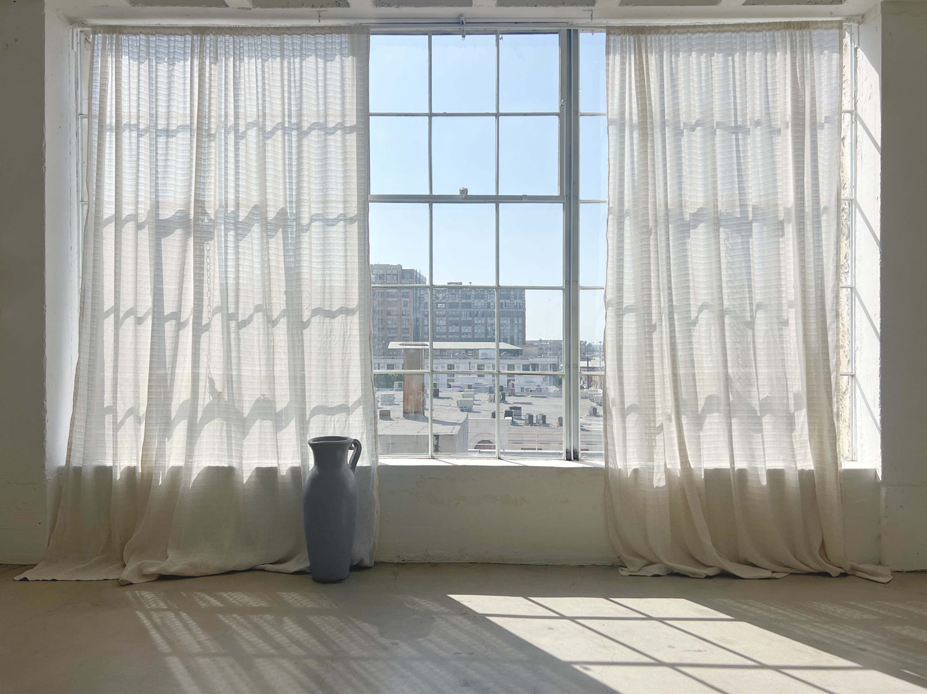 A large window with sheer white curtains frames a view of buildings and a paved area outside.