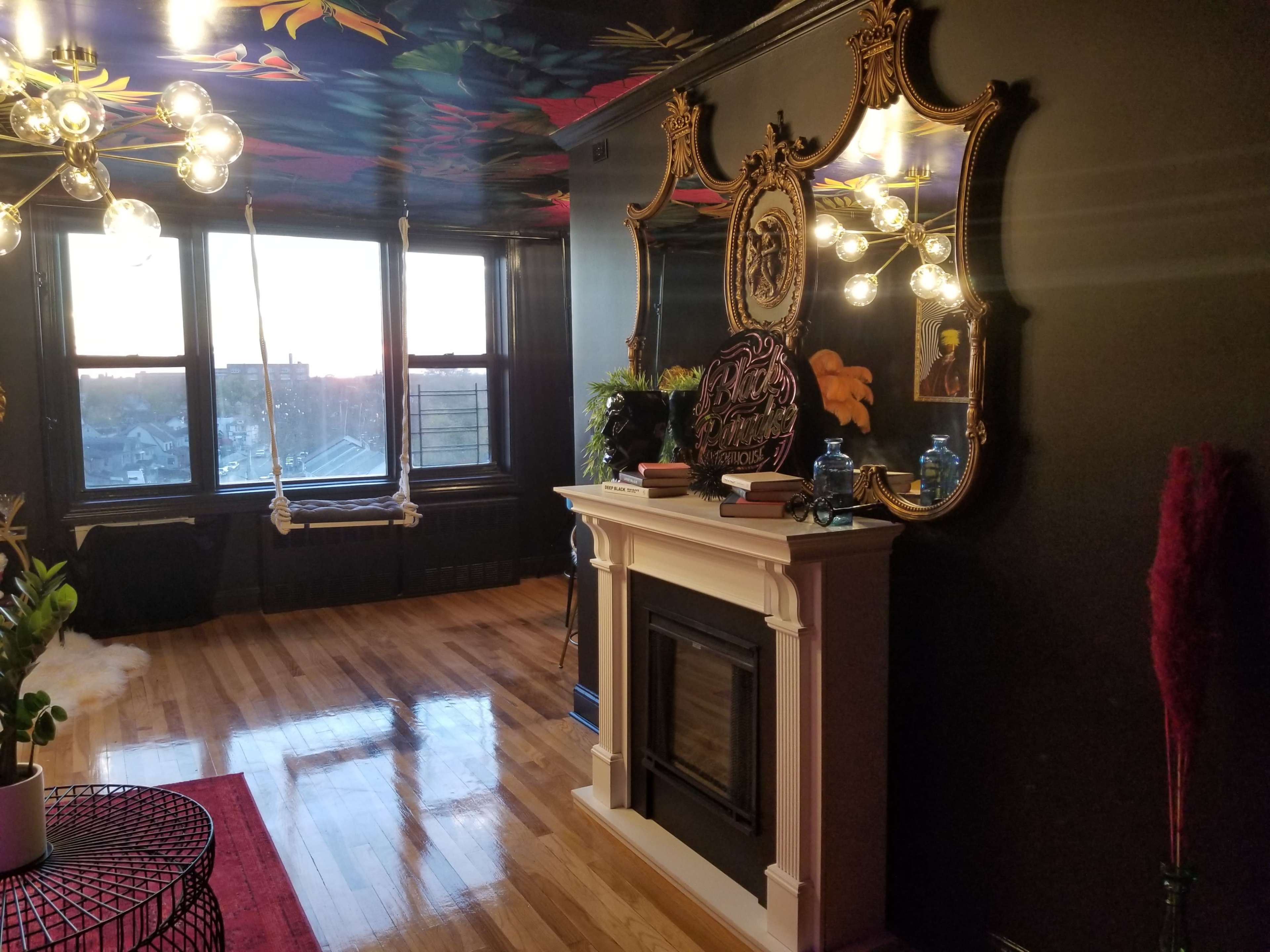 A cozy room features a fireplace surrounded by a decorative mirror, with large windows revealing a sunset view and a swing hanging from the ceiling.