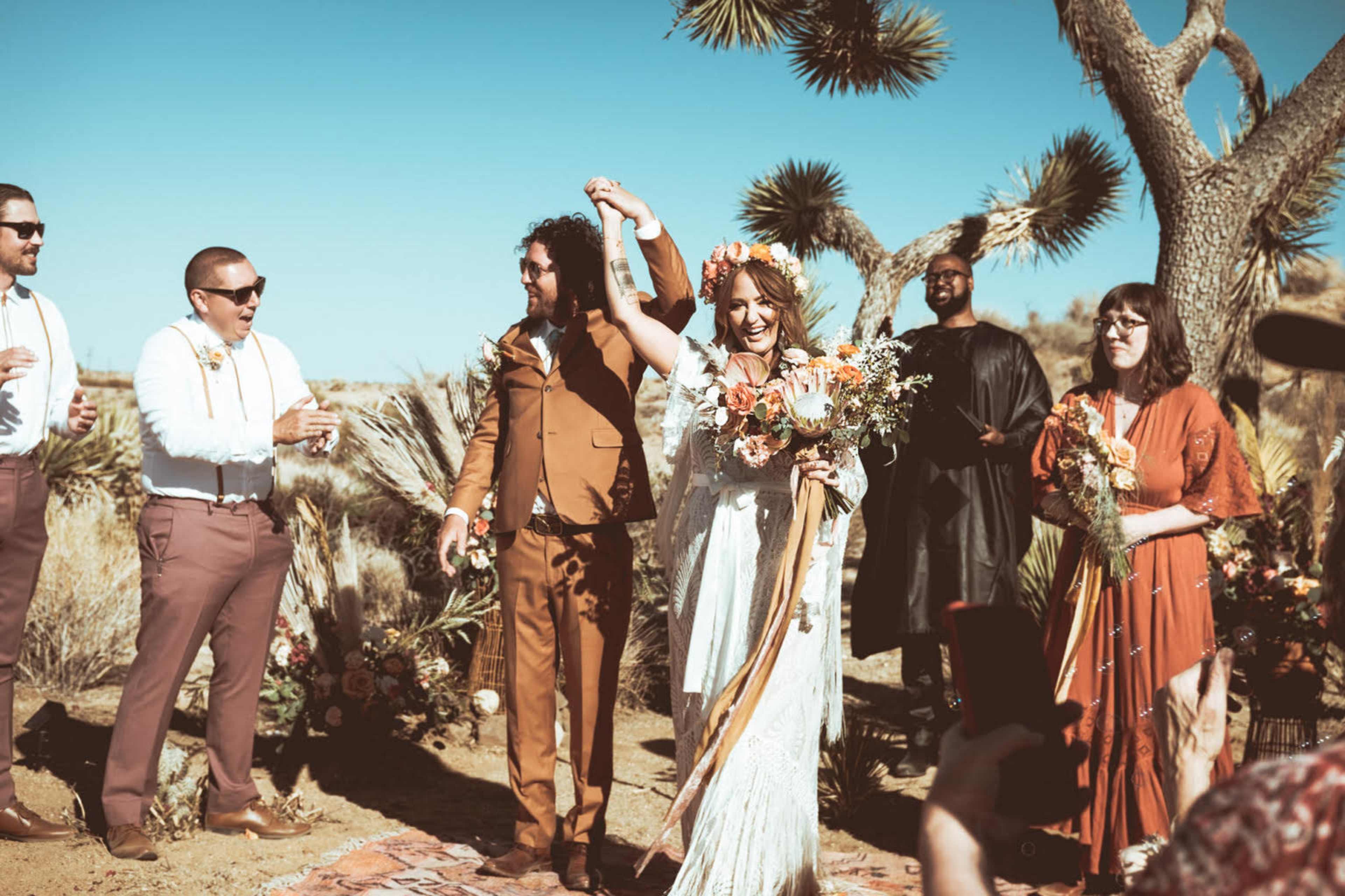 A couple celebrates their wedding under a clear blue sky in a desert setting, surrounded by guests holding flowers and drinks.