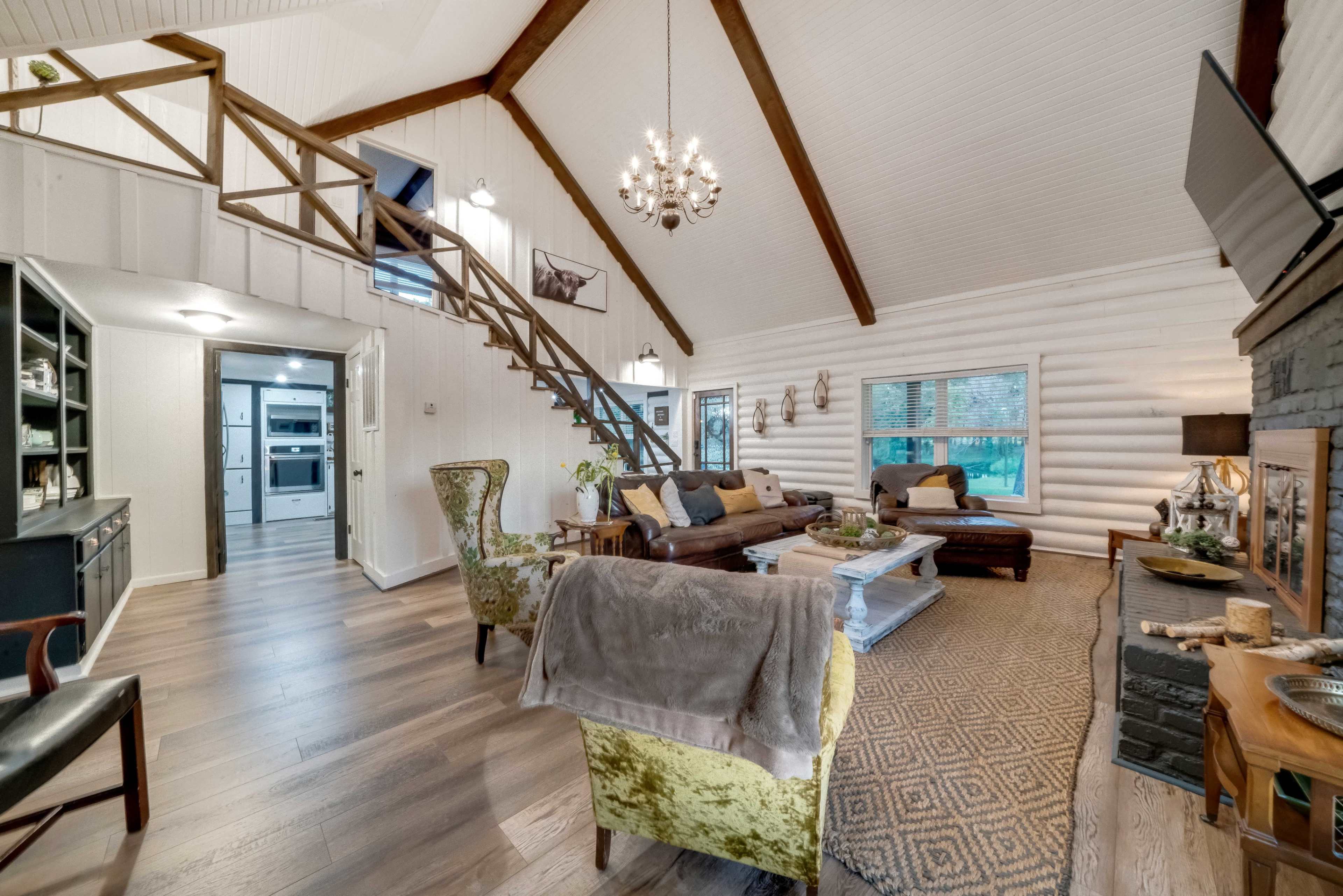 The image shows a spacious living room with a high ceiling, a staircase leading to a loft area, and a cozy seating arrangement around a central coffee table.
