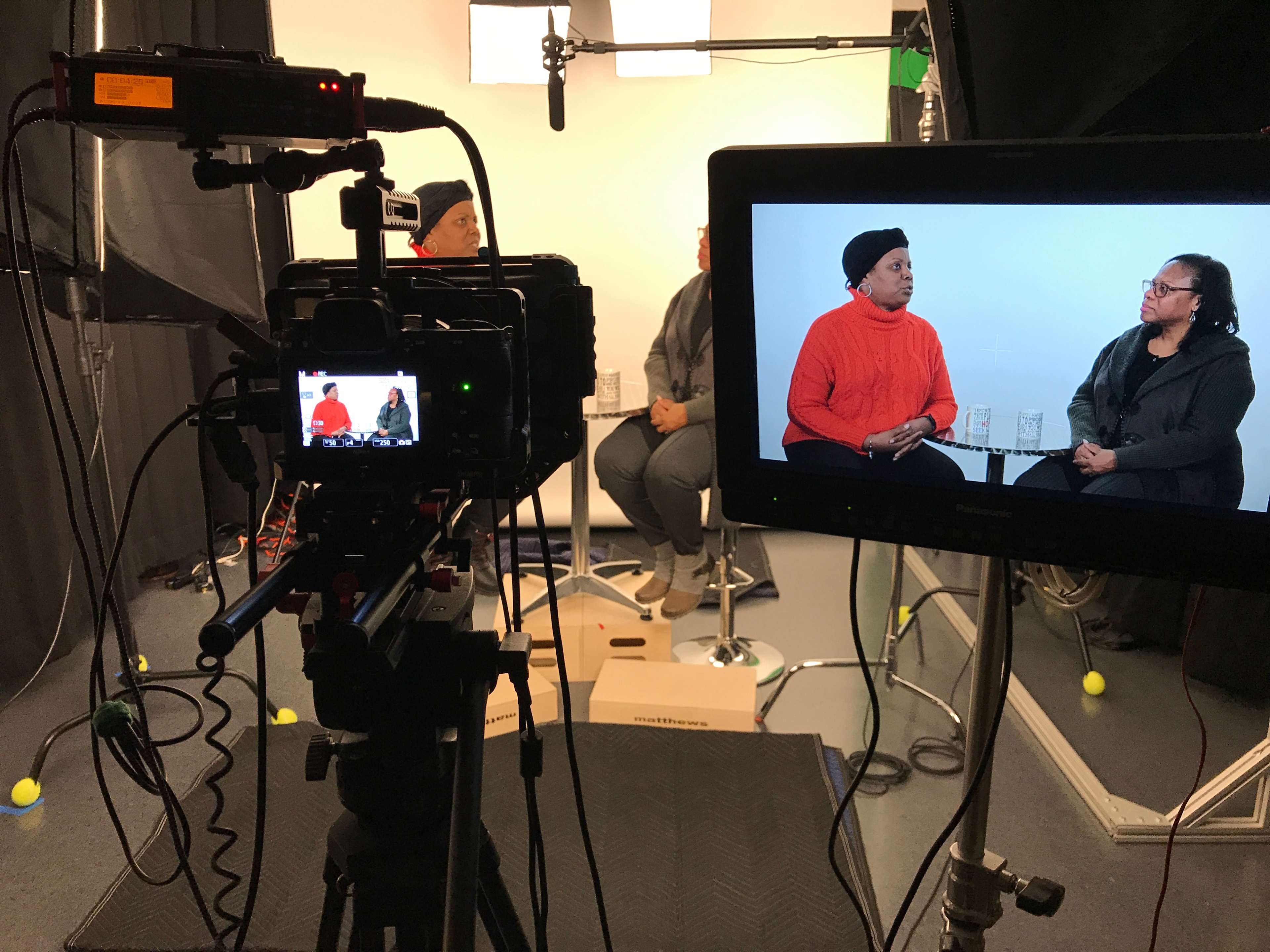 A video recording setup with two women seated for an interview, displayed on a monitor and captured by a camera in a studio environment.