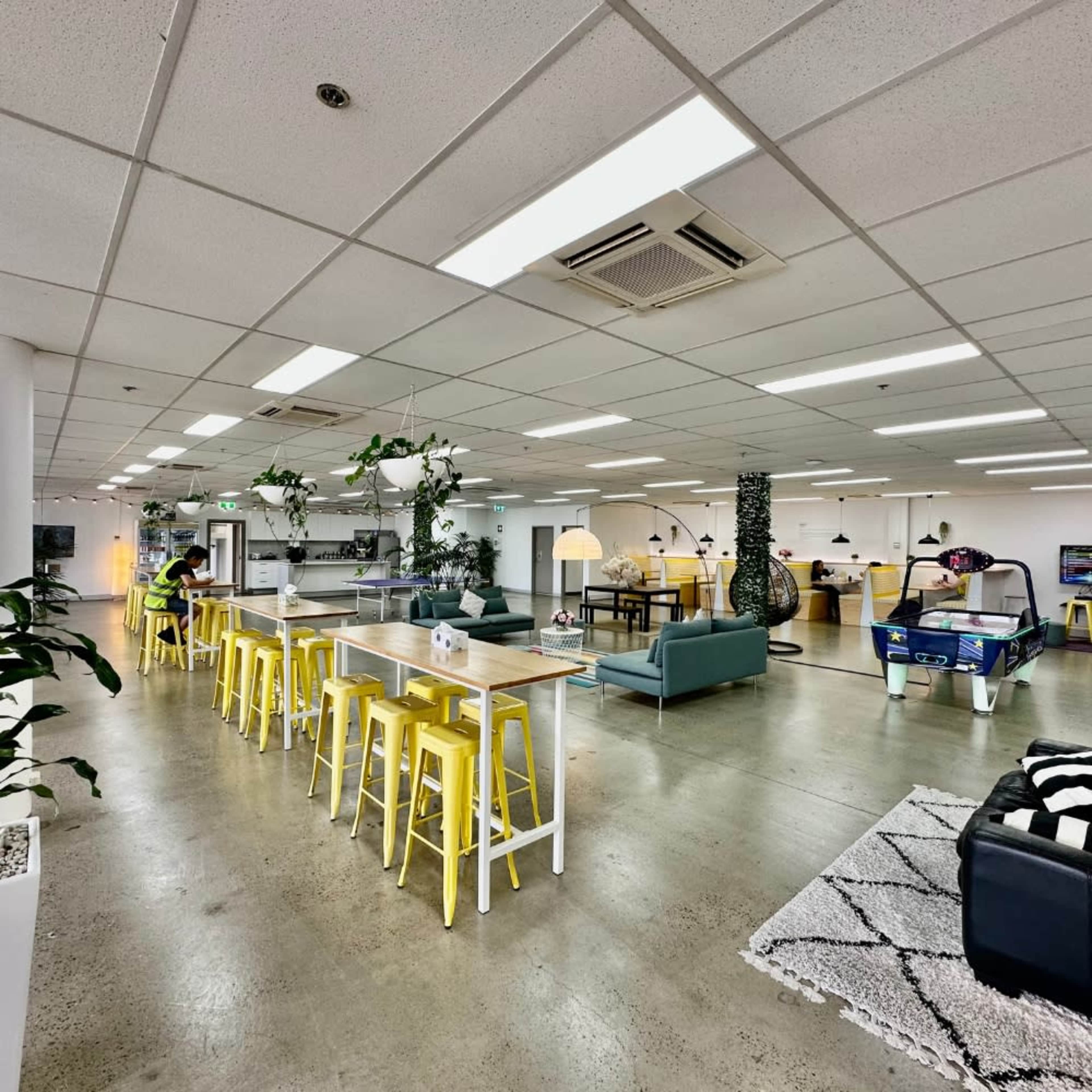 A modern office space with yellow stools at a long table, a lounge area with sofas, and a game table in a bright, open layout.