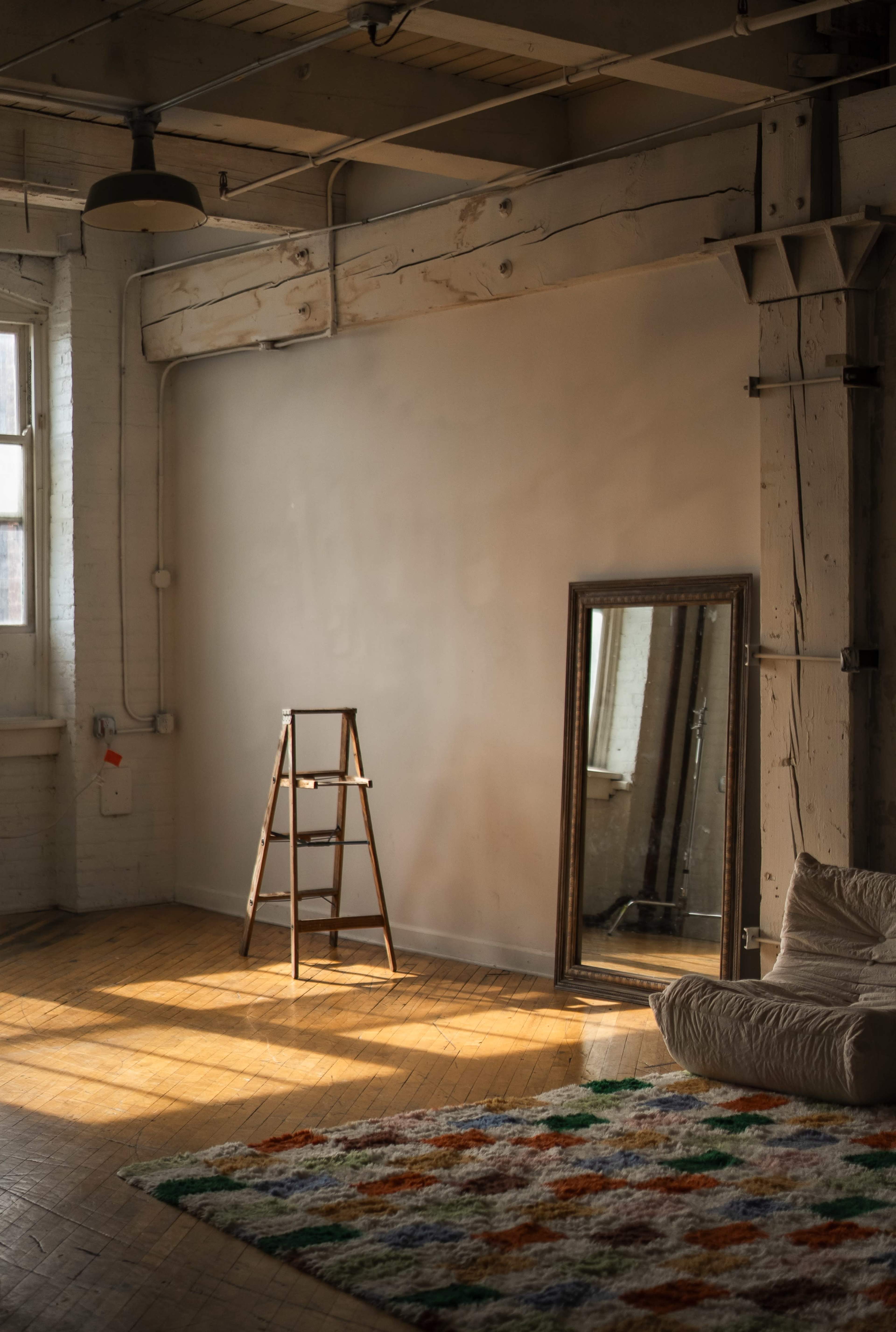 The image shows a spacious room with wooden floors, a leaning ladder against a bare wall, a large mirror reflecting light, and a colorful rug on the floor.