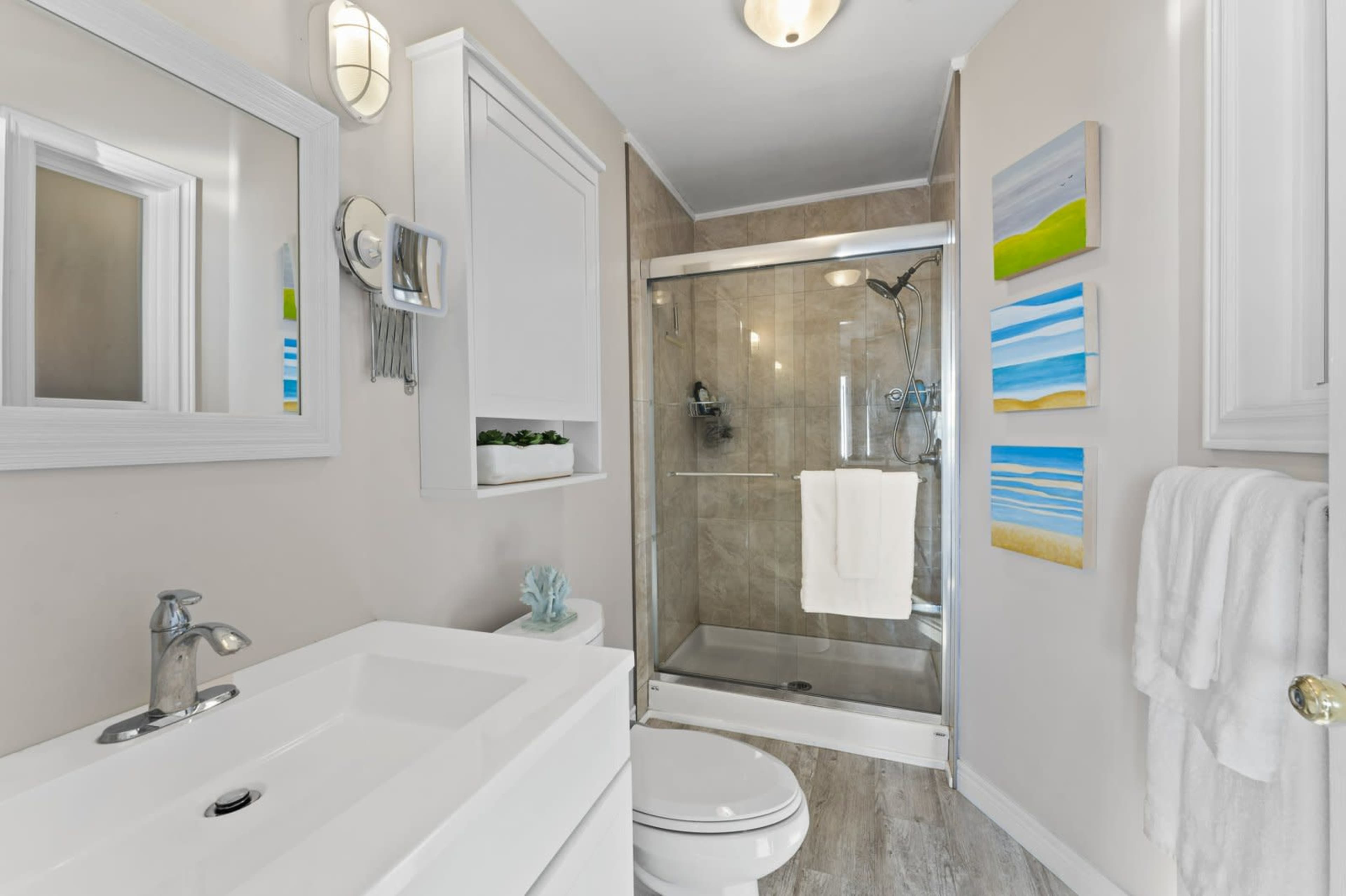The image shows a modern bathroom featuring a glass shower, a white vanity, and colorful artwork on the walls.